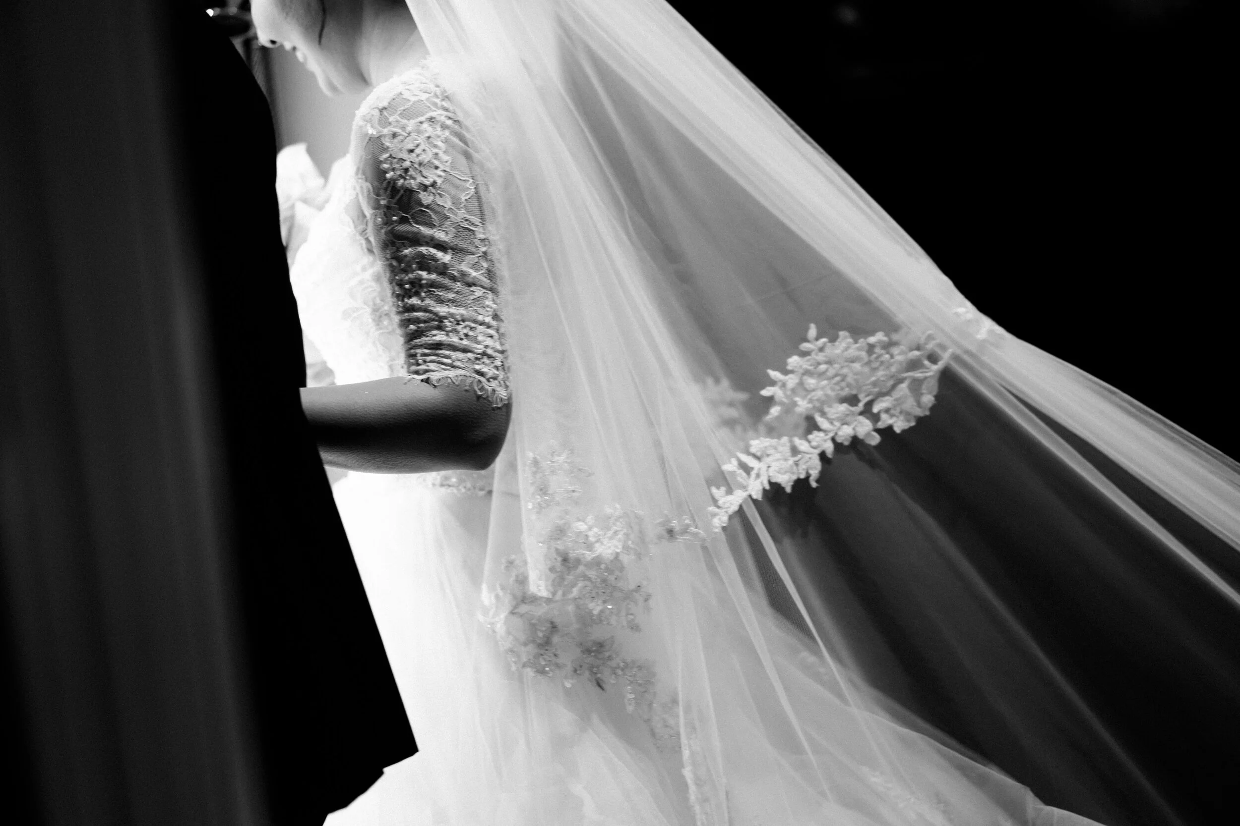 Close-up of a bride wearing a lace wedding dress with floral details and a long veil during a wedding ceremony.