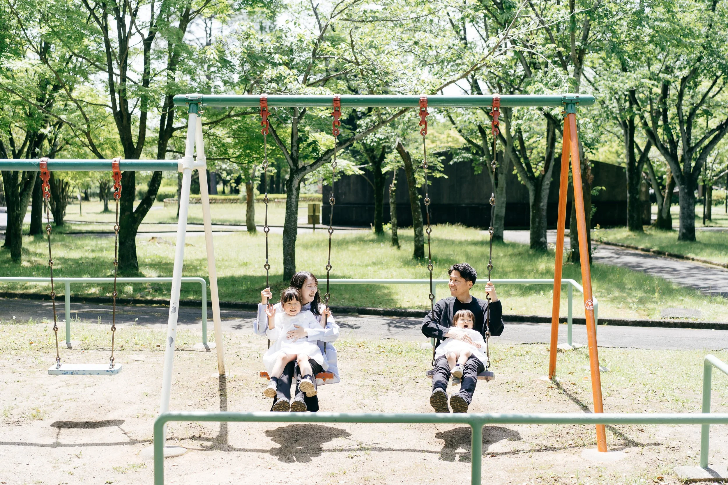A family of four enjoying time on swings at a park, with mature trees in the background and sunshine.