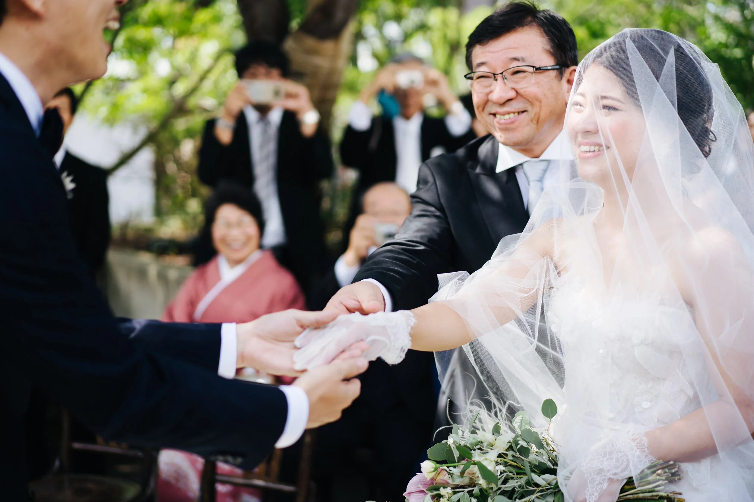 A bride and groom holding hands during an outdoor wedding ceremony, with guests taking photos in the background.