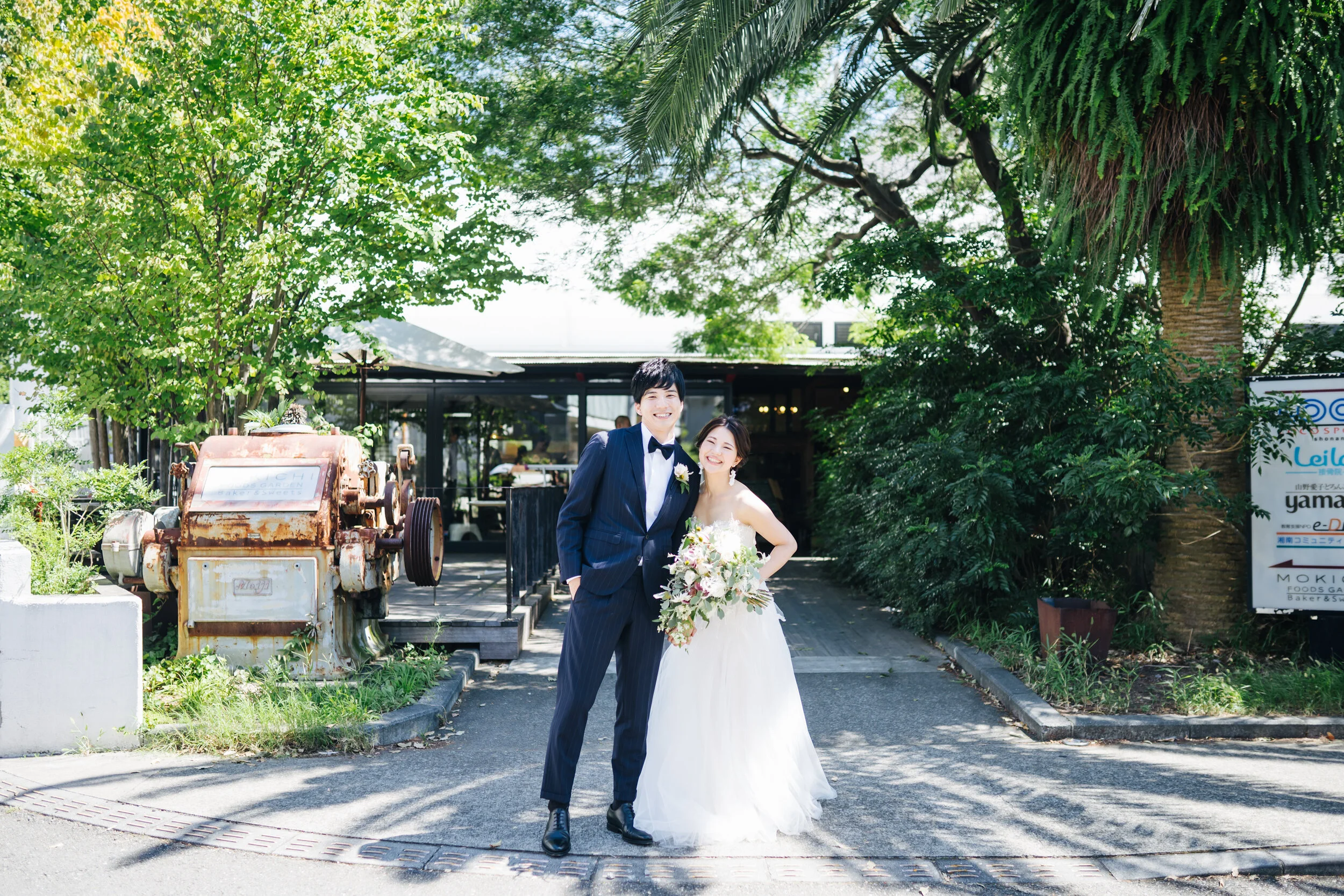 A bride and groom smiling outdoors, standing on a paved pathway under green trees, with the bride holding a large bouquet of flowers.