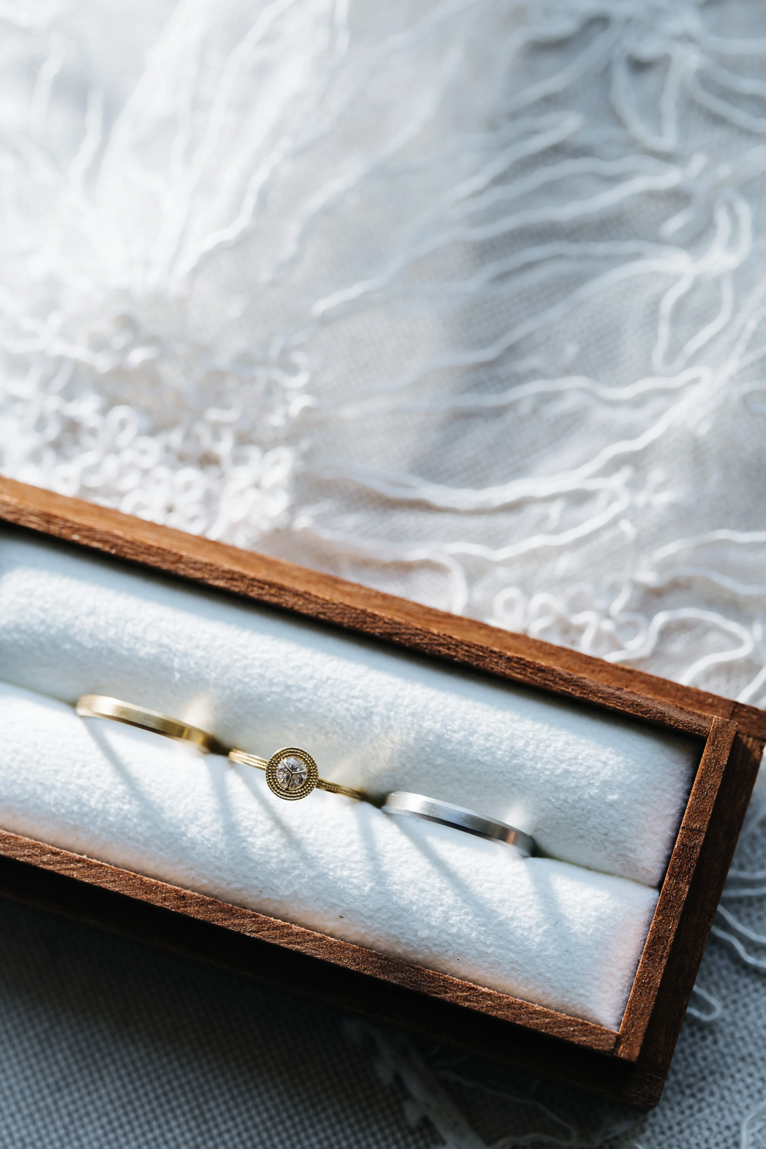 A jewelry box with a white velvet interior contains two rings, one with a central diamond surrounded by smaller stones and the other is a plain band, placed on a dark surface with a piece of fabric nearby.