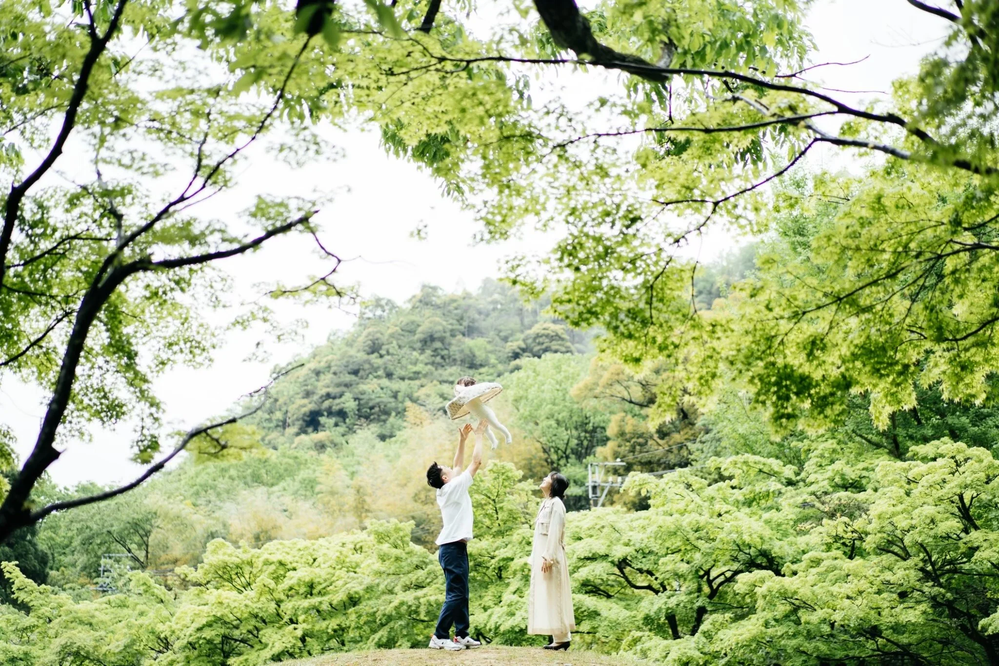 A couple with the man lifting a child dressed in white in a lush green forest with two women standing nearby, under a canopy of green leaves.