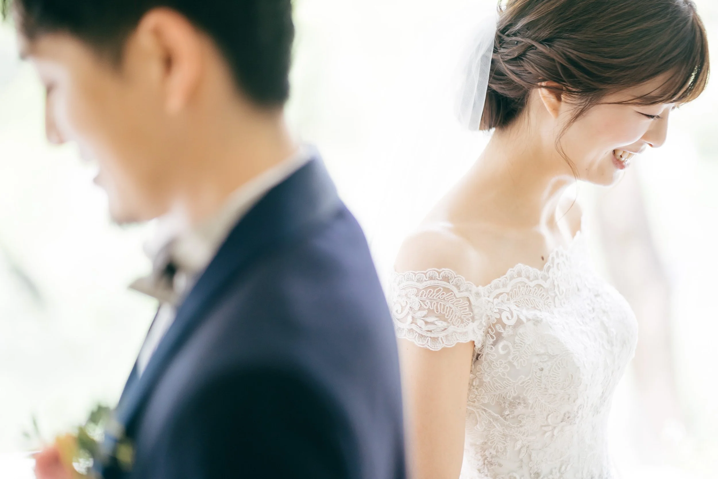 A bride and groom at their wedding, both smiling and looking down, with the bride wearing a lace wedding dress and the groom in a suit.