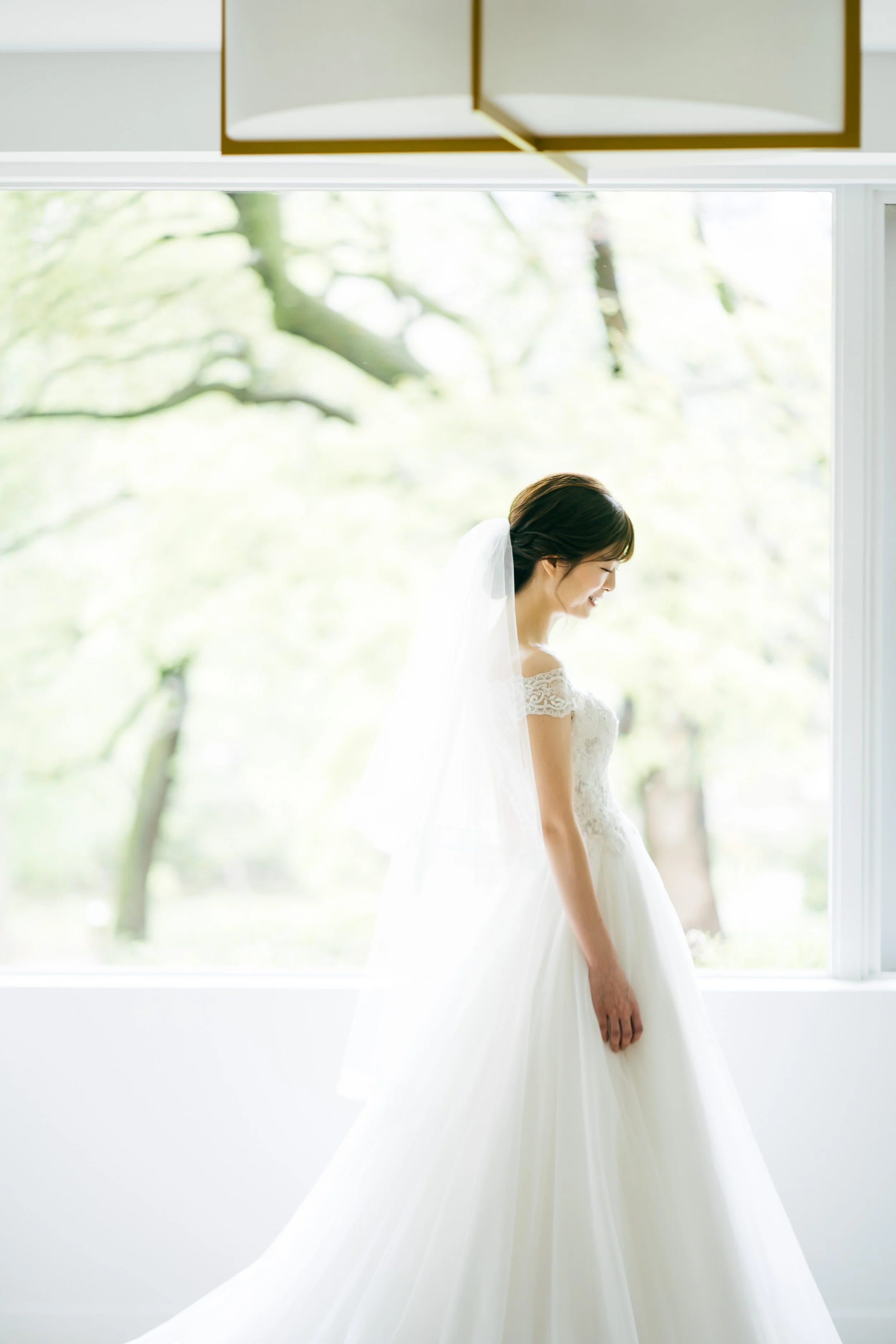 A bride in a white lace wedding dress and veil standing near a large window with a view of greenery outside, smiling.