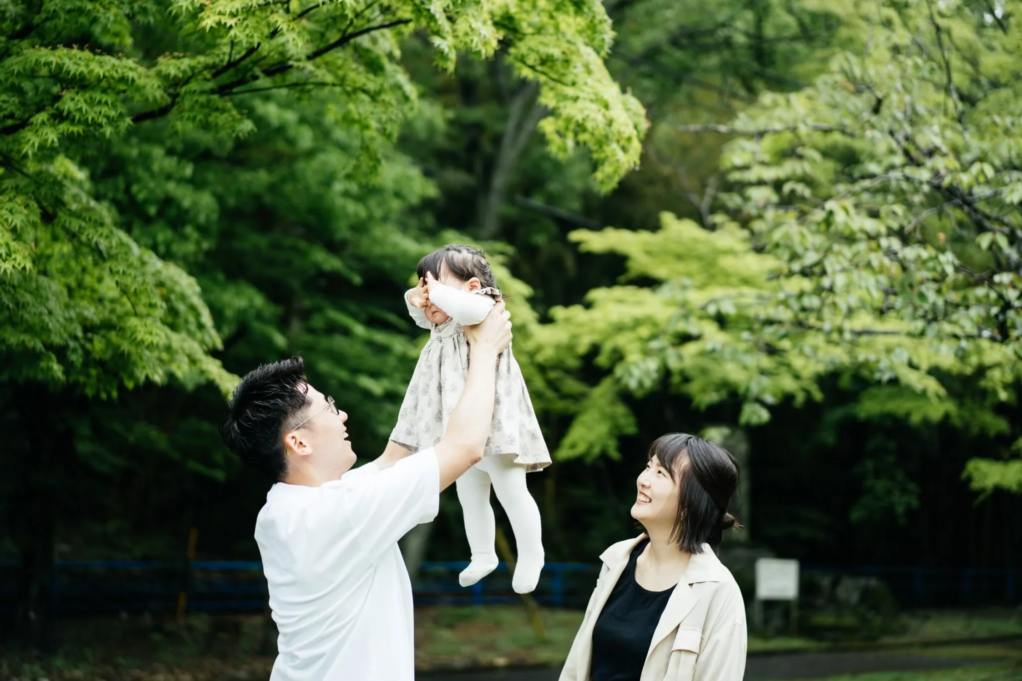 A man lifts a young girl into the air in a park surrounded by green trees. The young girl has her hands covering her face and wears a light-colored dress. A woman with black hair and a tan blazer looks up at the girl, smiling. Another person partiall
