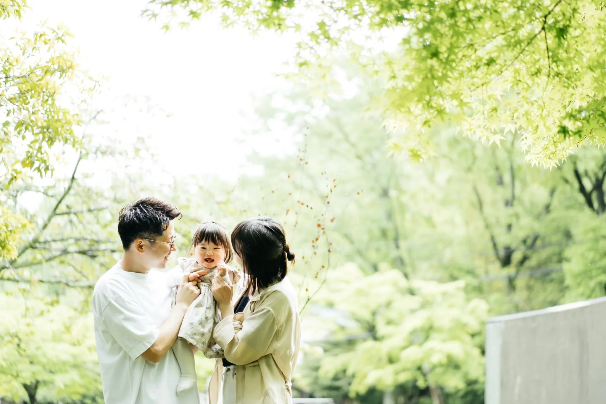 A family of three holding a young child outdoors in a lush green park with trees overhead, as they enjoy a sunny day.