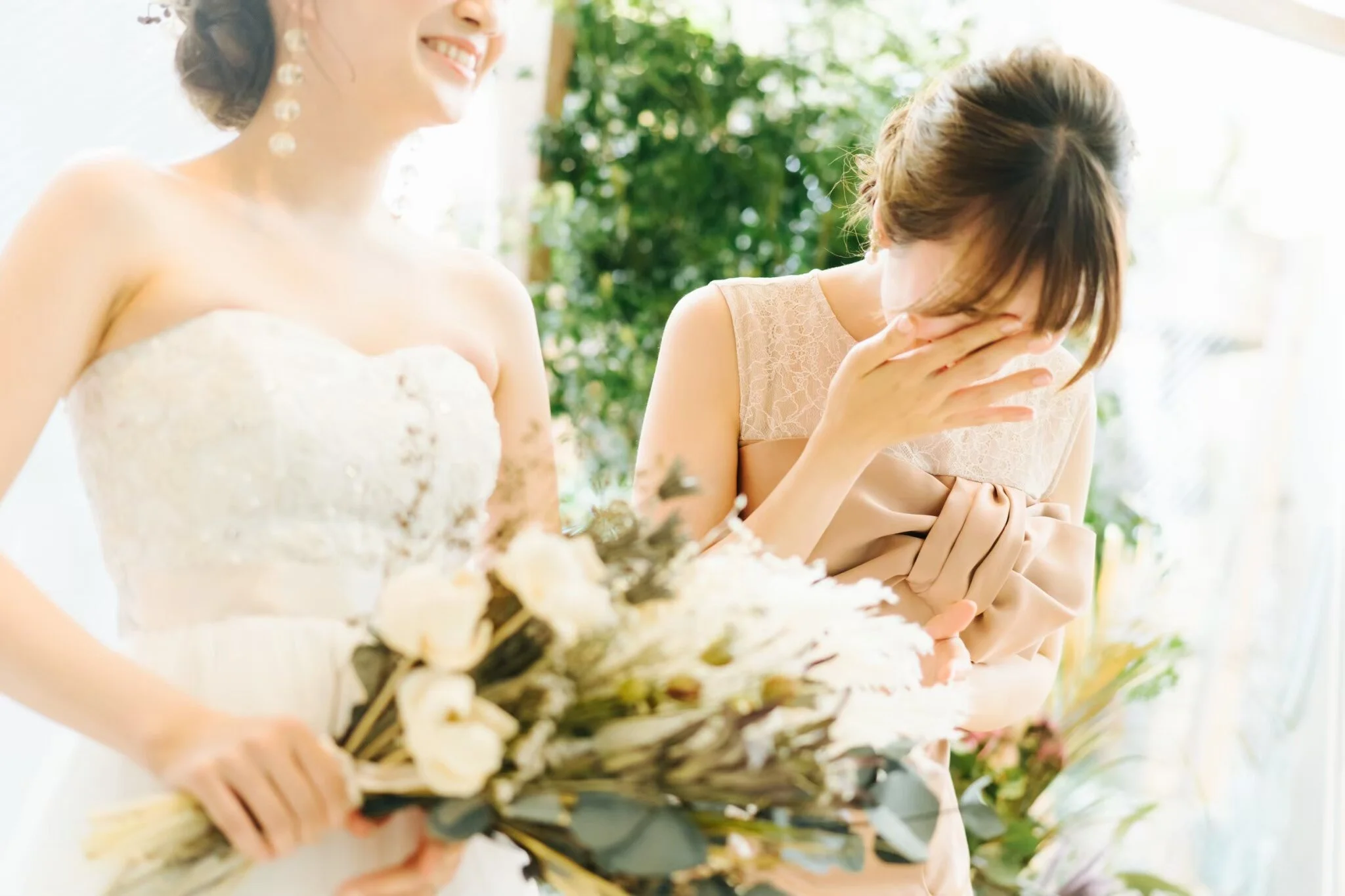 A bride in a white wedding dress smiling and standing next to a woman covering her face with her hand, possibly emotional, with greenery in the background.