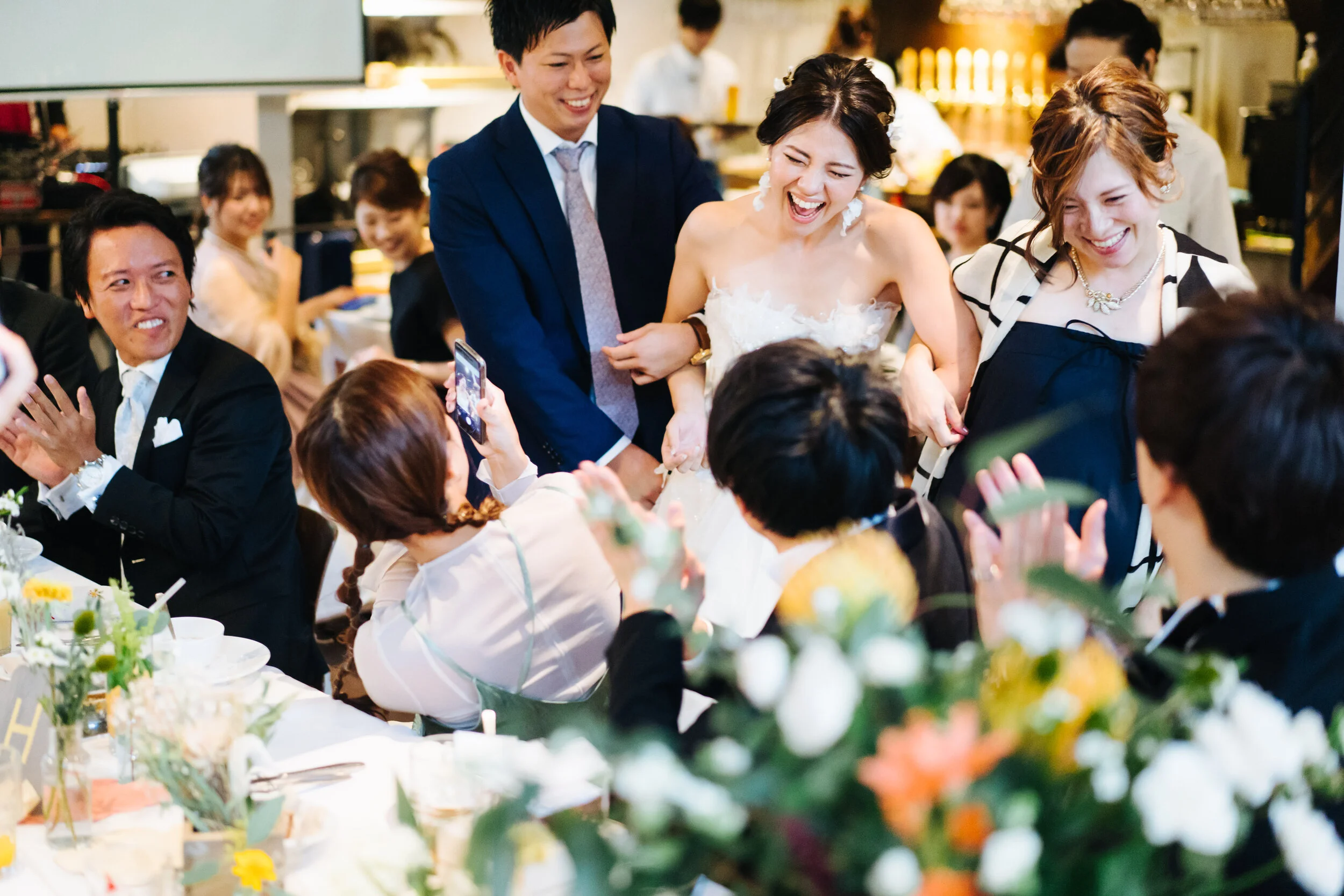 People smiling and laughing at a wedding reception, with a bride in a white dress, guests in formal attire, and floral centerpieces on tables.