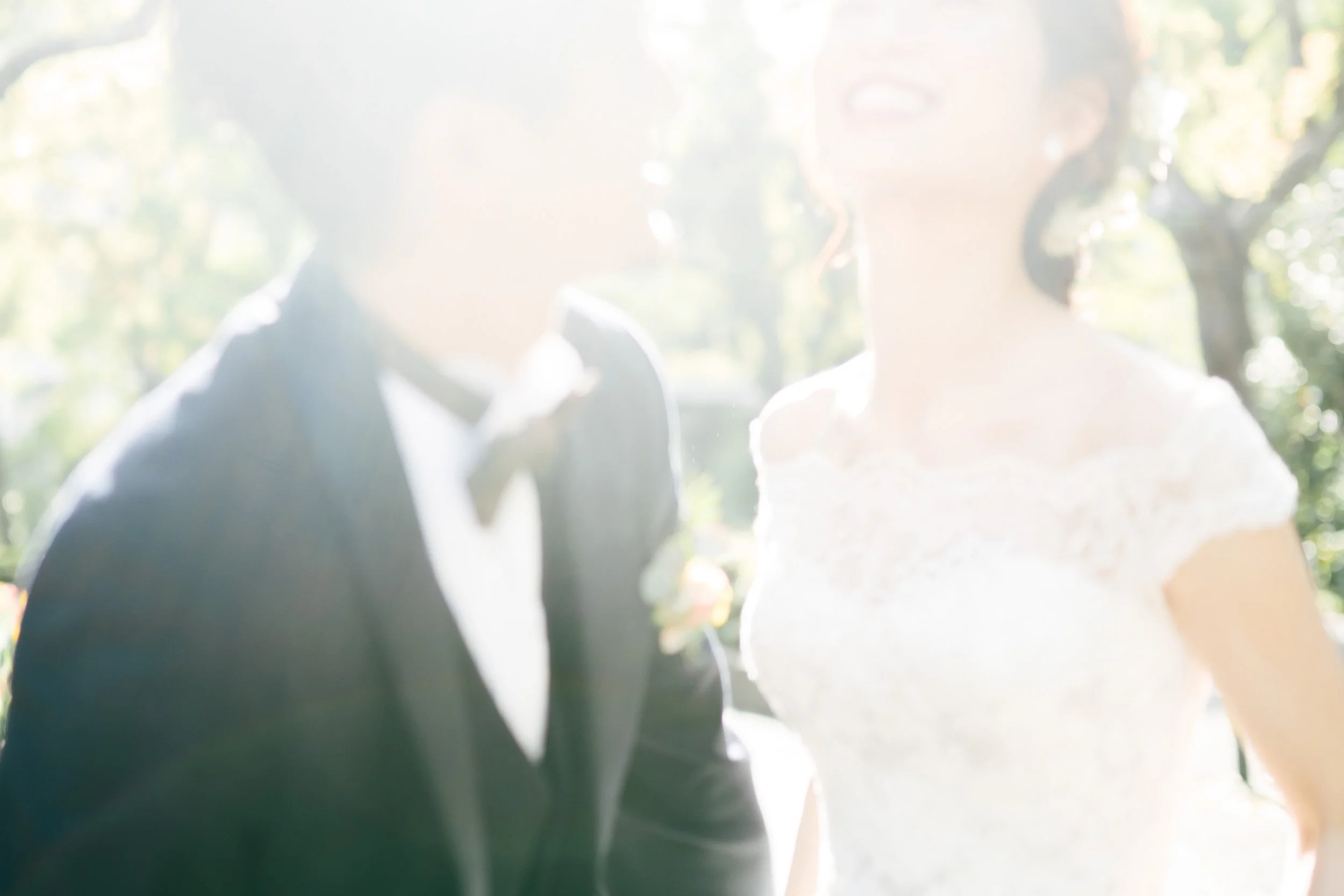 A blurry photo of a bride and groom outdoors, with sunlight filtering through trees in the background.