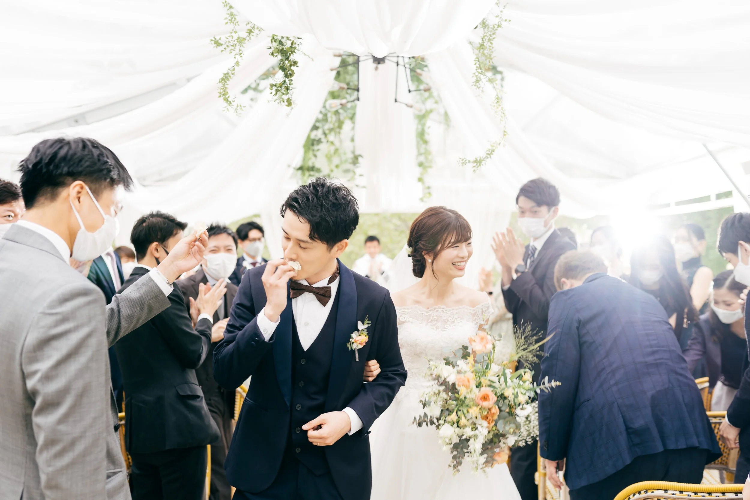 A wedding celebration where a groom in a navy suit and a bride in a white lace dress are smiling and holding hands, surrounded by guests wearing masks under a white draped canopy with hanging greenery.