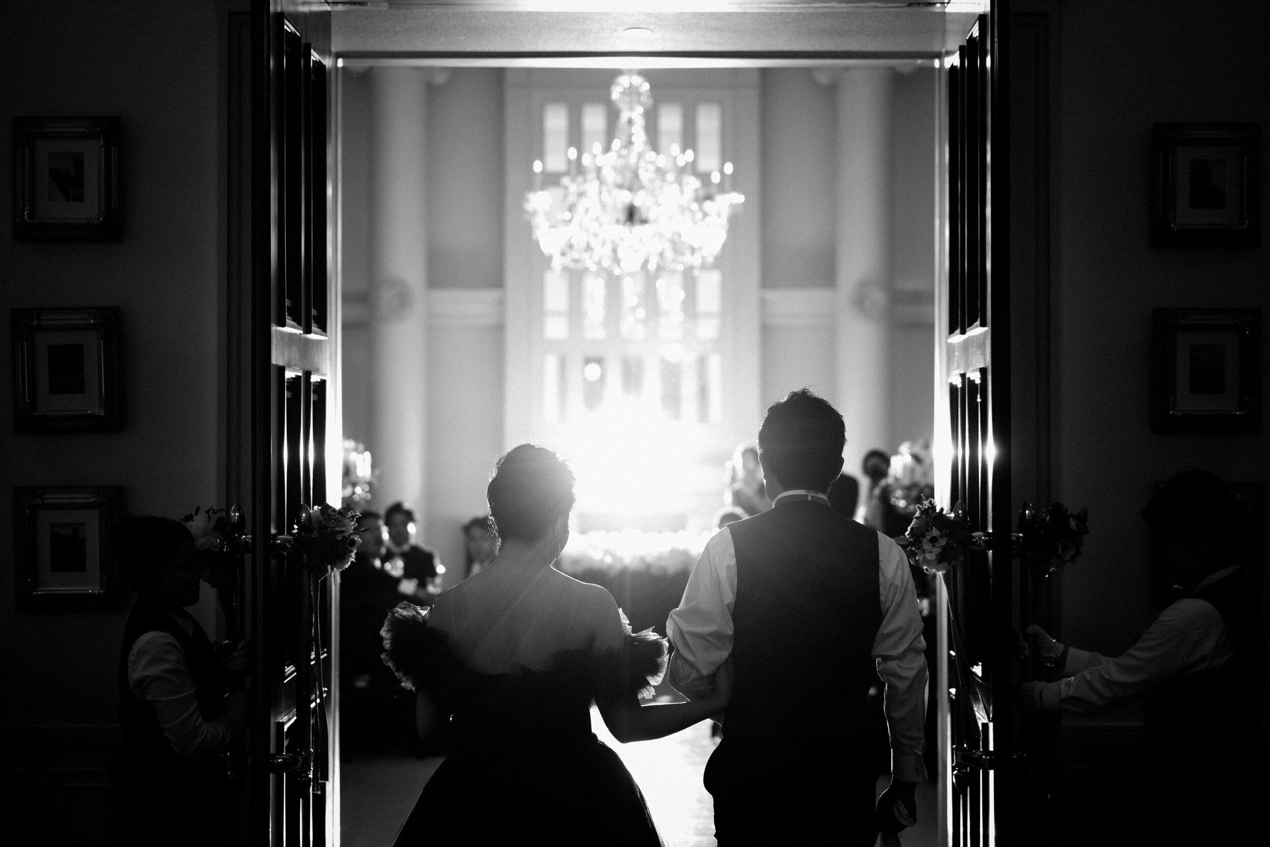 Silhouettes of a bride and groom holding hands, walking through open doors into a brightly lit room with a large chandelier in the background, during a wedding ceremony.