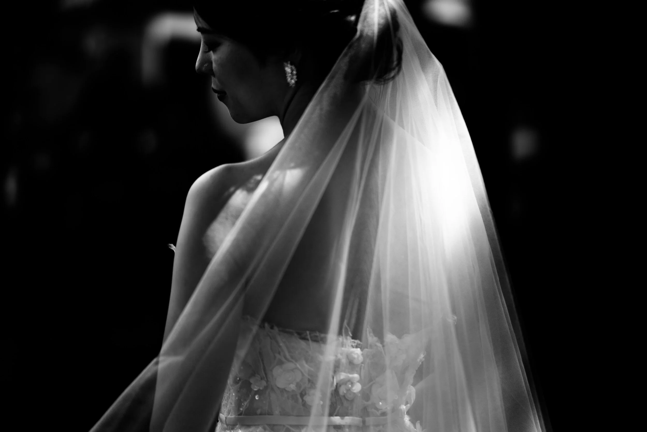Black and white photograph of a bride with her head bowed, wearing a veil and pearl earrings, with soft lighting highlighting her profile.