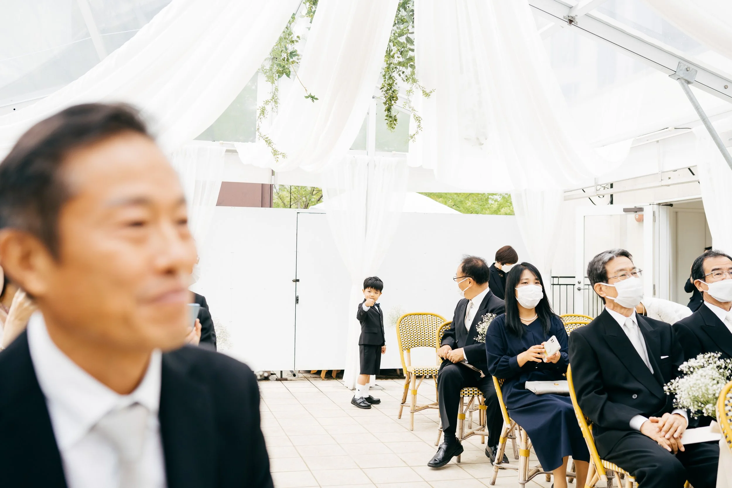 People attending an outdoor event, seated in chairs wearing formal attire and face masks, with a white tent and hanging greenery overhead.