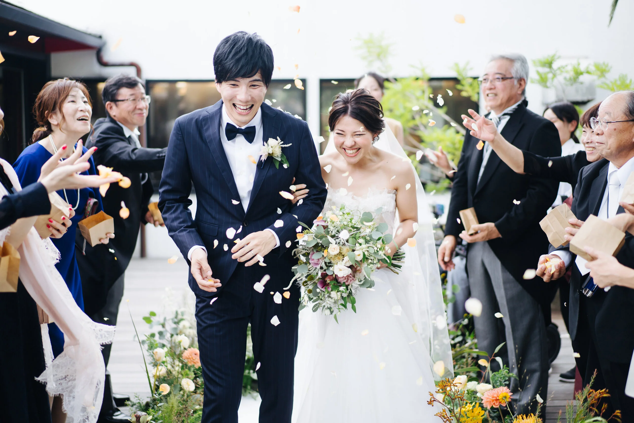 A group of wedding guests celebrating outdoors as a bride and groom walk through, tossing flower petals, with happy expressions and spring floral decorations.