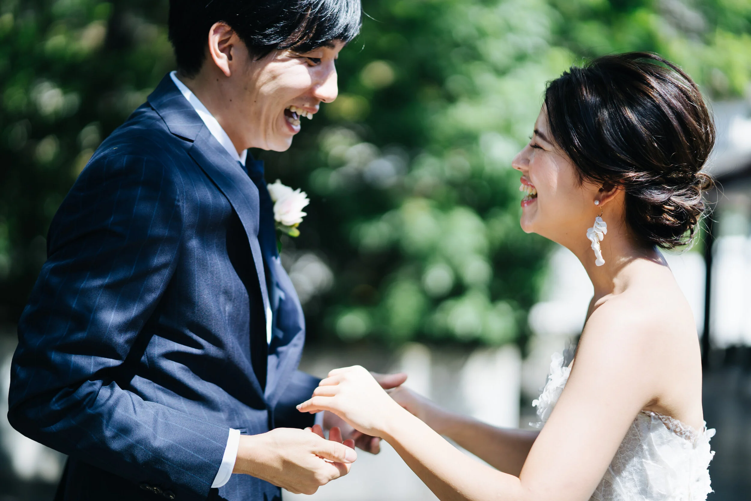 A couple on their wedding day, holding hands, smiling, outdoors with green foliage in the background.