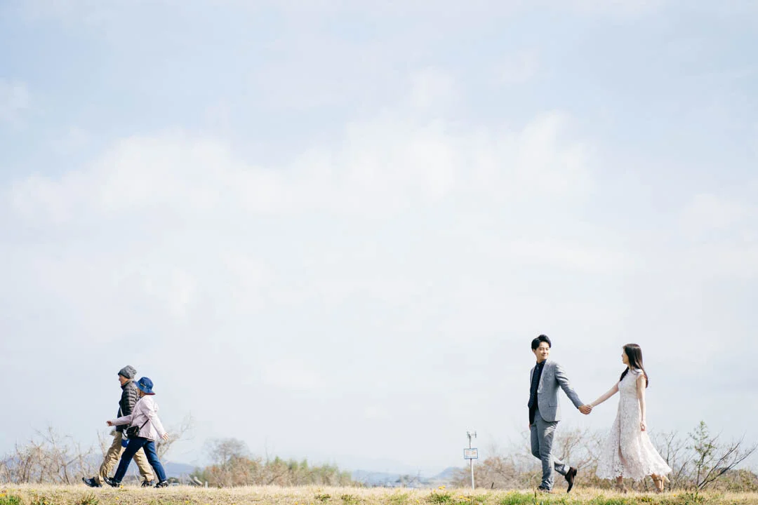 A man and woman holding hands outdoors, walking on a grassy field, with other people walking in the background.
