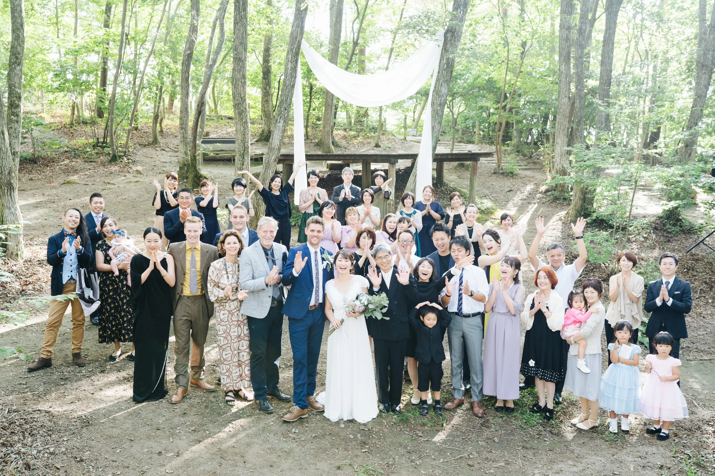 A large group of people, including children and adults, gathered outdoors in a wooded area for a wedding celebration. The bride and groom are at the front, smiling and waving. Trees and greenery surround the group, and a white cloth drapes between tw