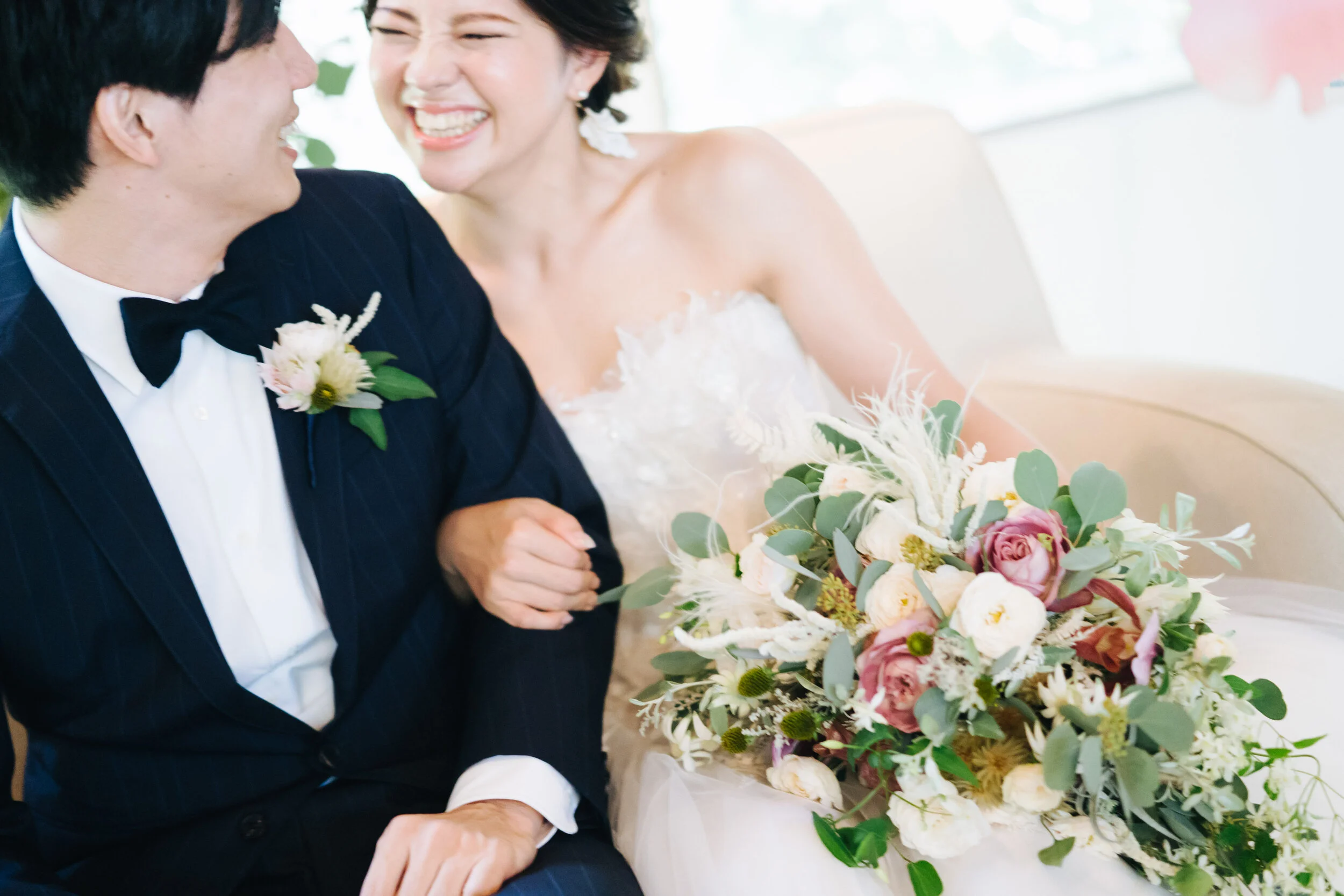 A smiling bride and groom holding hands, sitting next to a large bouquet of flowers on their wedding day.