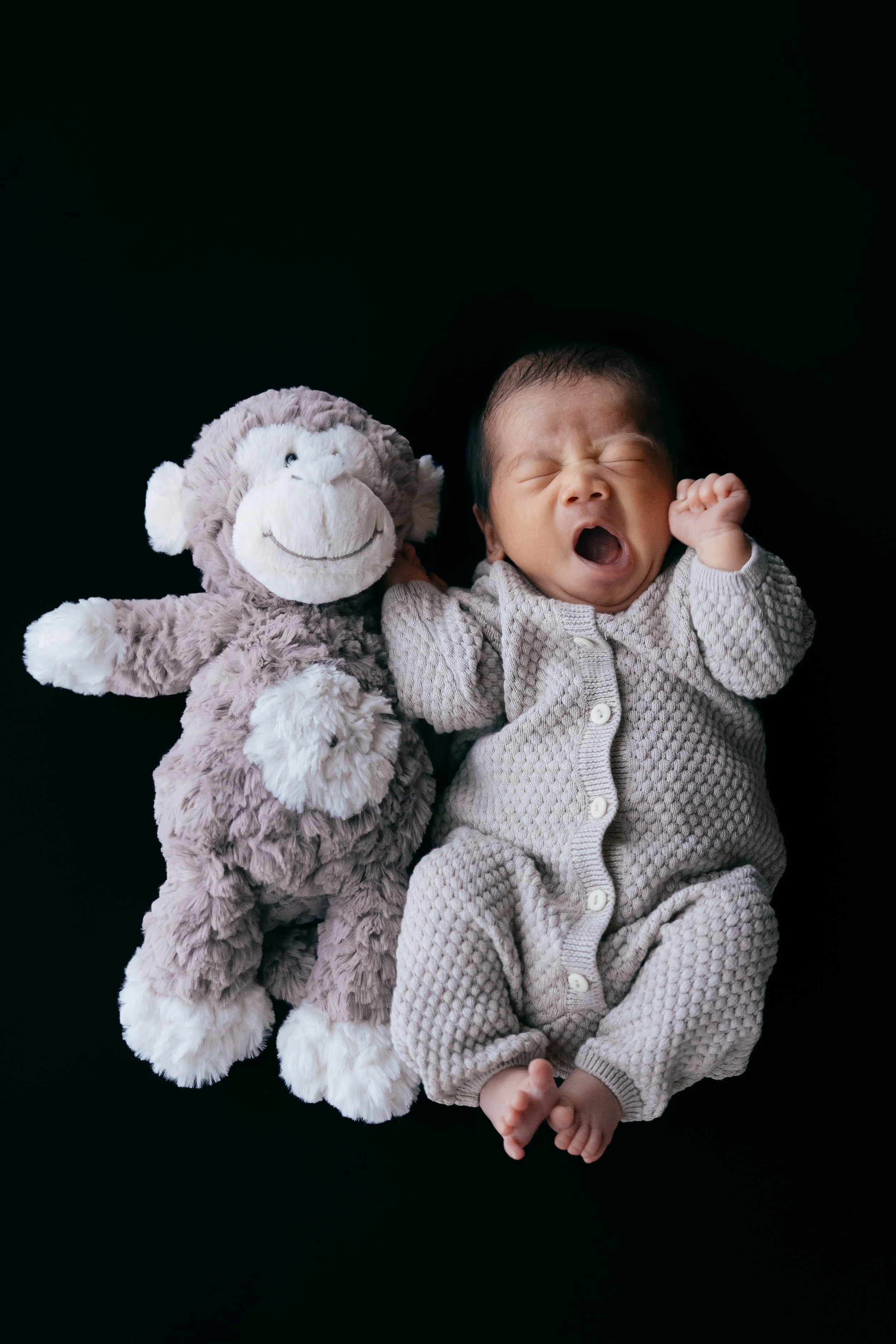 A newborn baby with closed eyes and an open mouth yawning, lying next to a plush monkey toy on a black background.