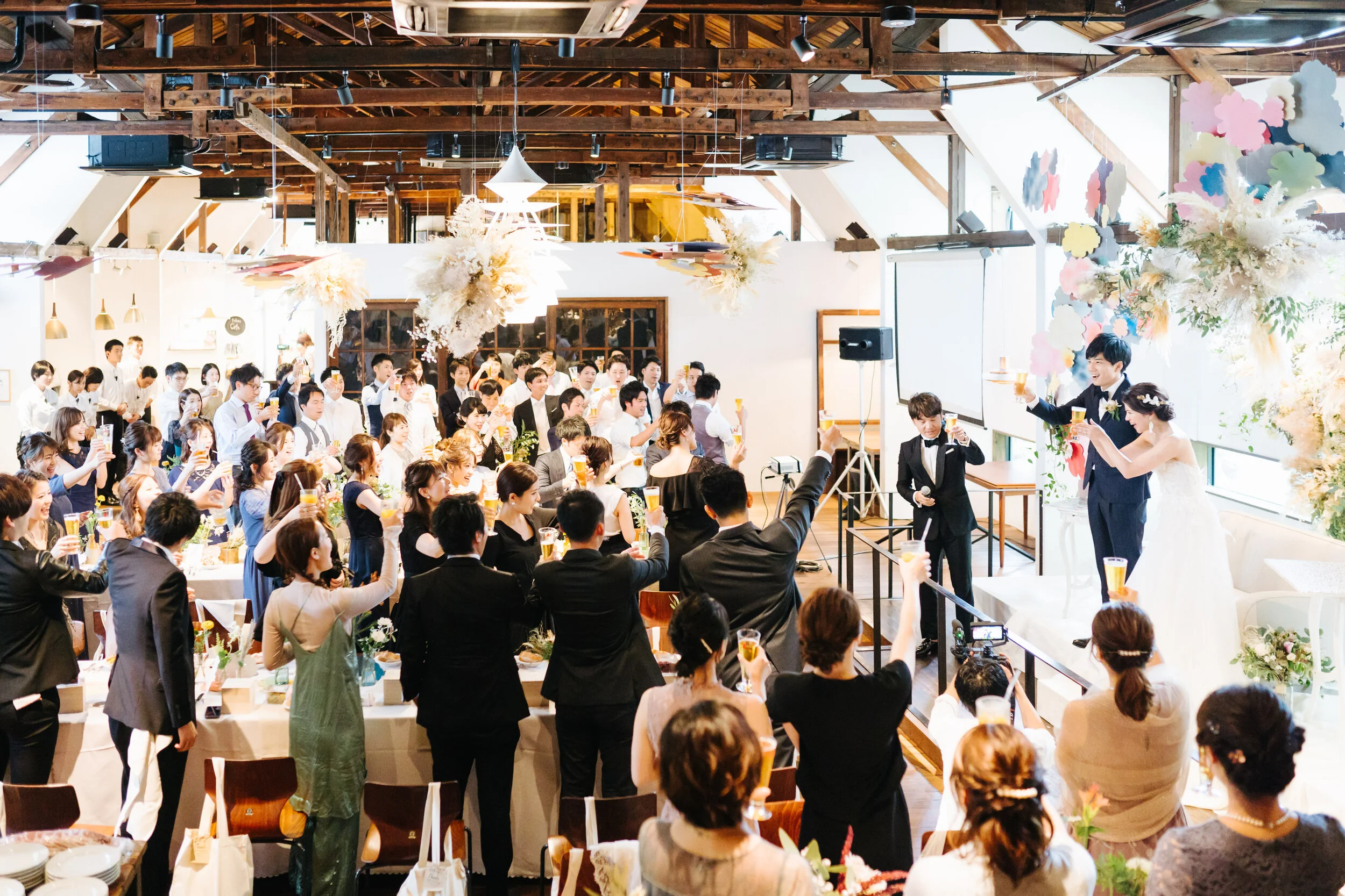 Wedding reception with bride and groom raising glasses, surrounded by guests in an airy, wood-beamed decorated hall with floral arrangements and pastel-colored wall decorations.