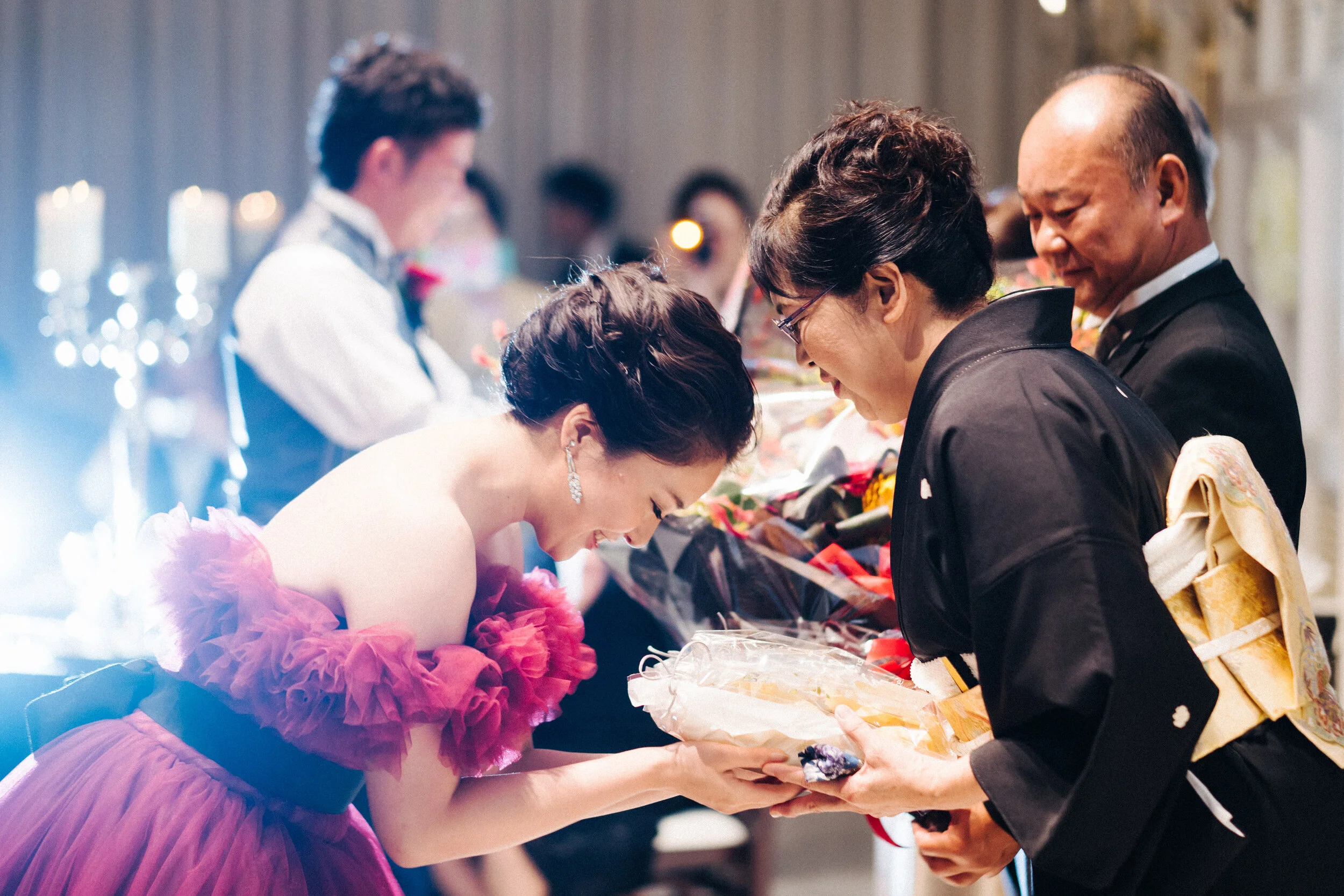 A young woman in a pink dress bows her head while receiving a handshake and a gift from an older woman and man at a formal event, with other people blurred in the background.