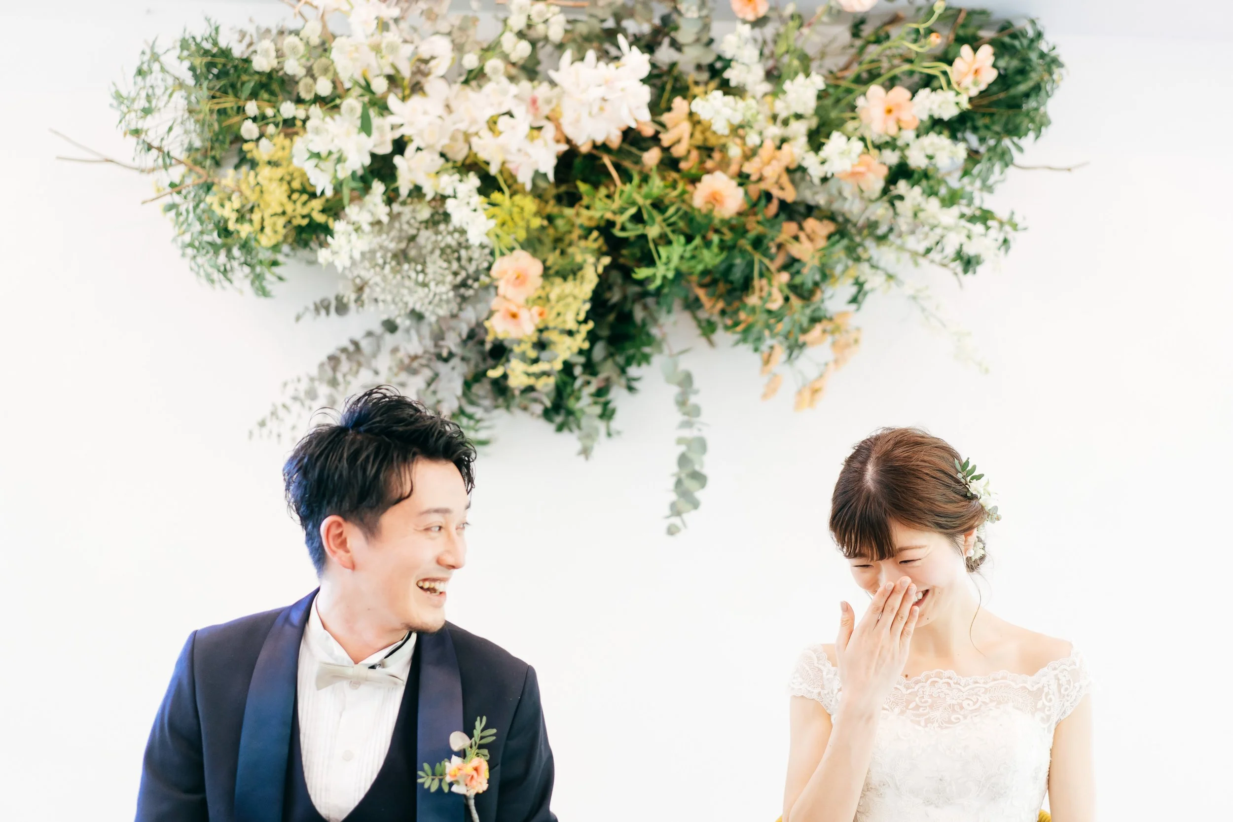 A couple dressed in wedding attire, smiling and laughing during their wedding ceremony. There is a large floral arrangement above them with white and blush flowers and green foliage.