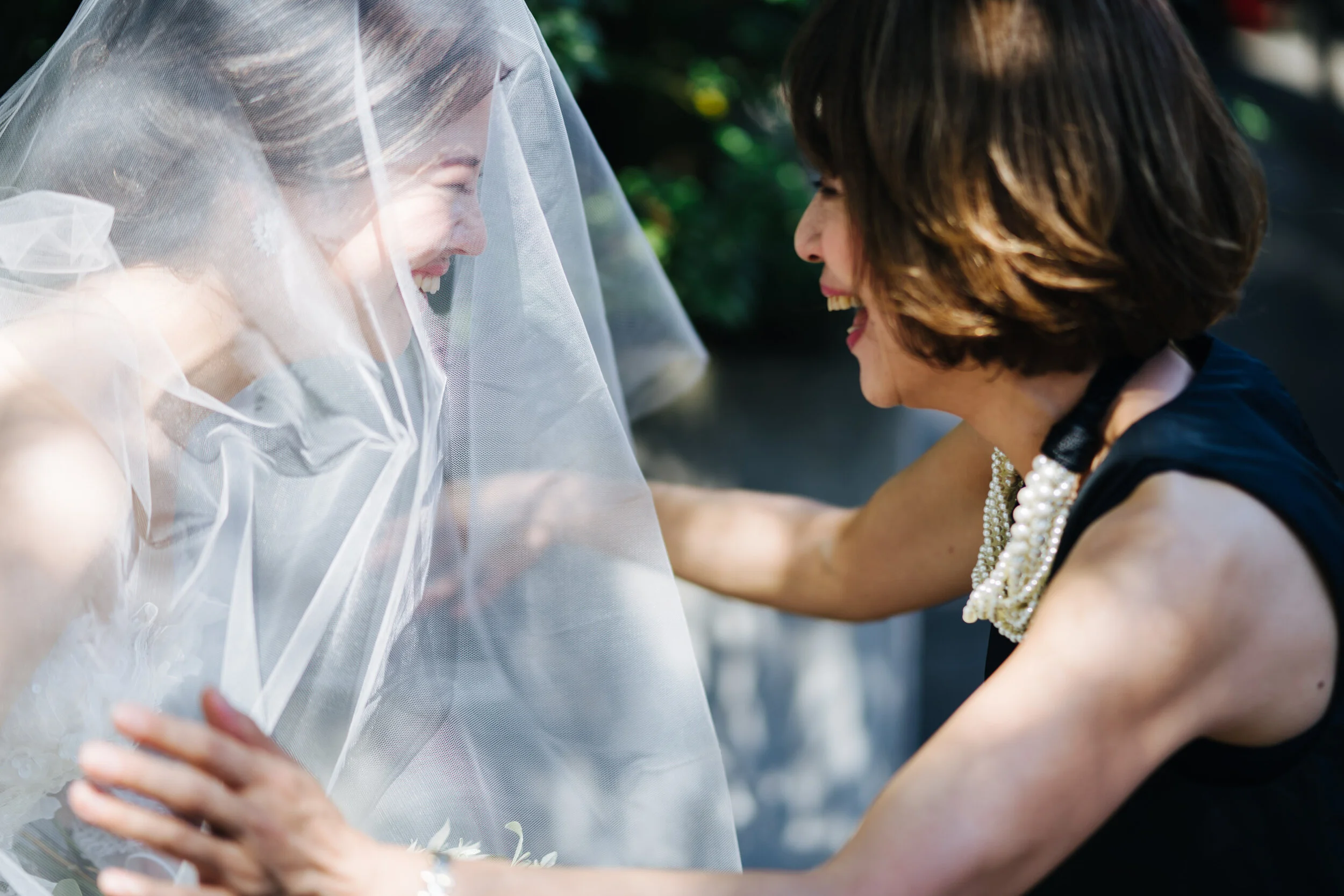 A bride with a veil and a woman smiling and laughing in an outdoor setting with greenery.