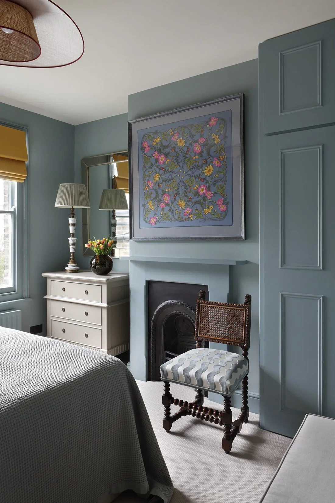 A cozy bedroom with a blue wall, a white dresser with a flower vase and table lamps, a framed floral artwork above a fireplace, a striped upholstered chair, a bed with a textured blanket, and yellow window shades.