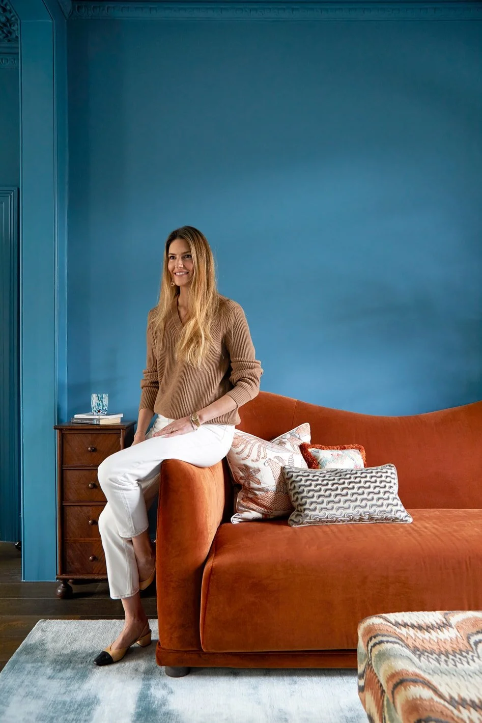 A woman with long blonde hair sitting on the arm of a burnt orange velvet sofa, smiling, in a room with a vibrant blue wall and wooden furniture, decorated with patterned pillows and a glass of water on a small bedside table.