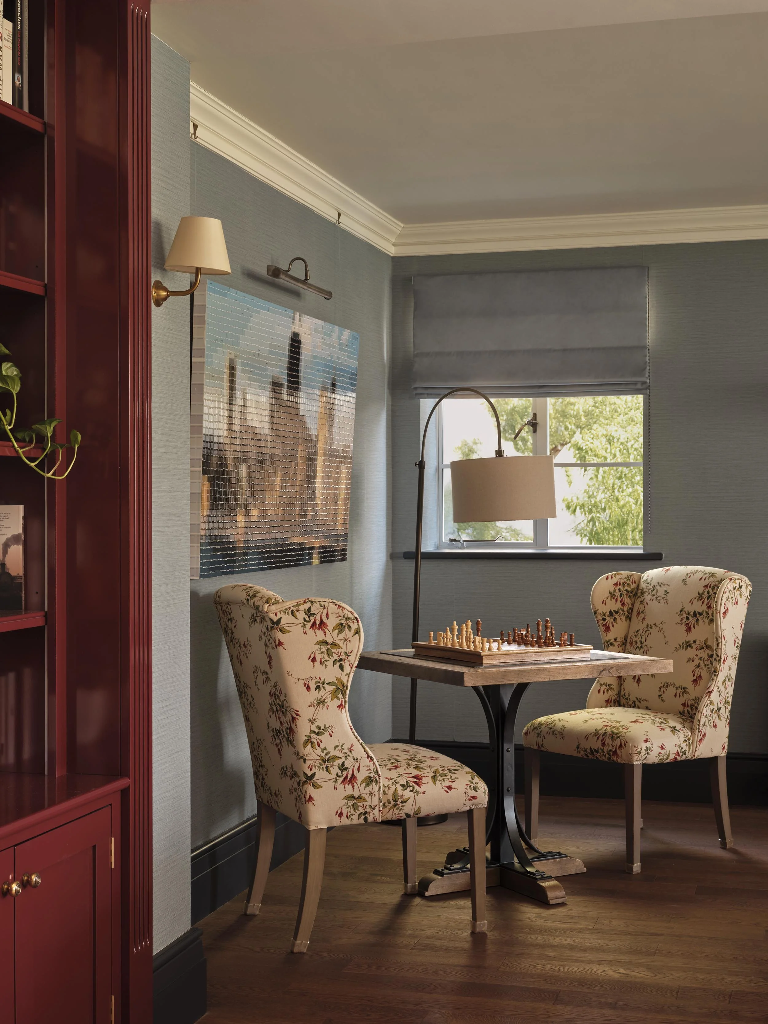 A cozy room with a chessboard on a small round table, surrounded by two floral upholstered chairs, near a window with a gray blind, and a large colorful city's skyline artwork on the wall.