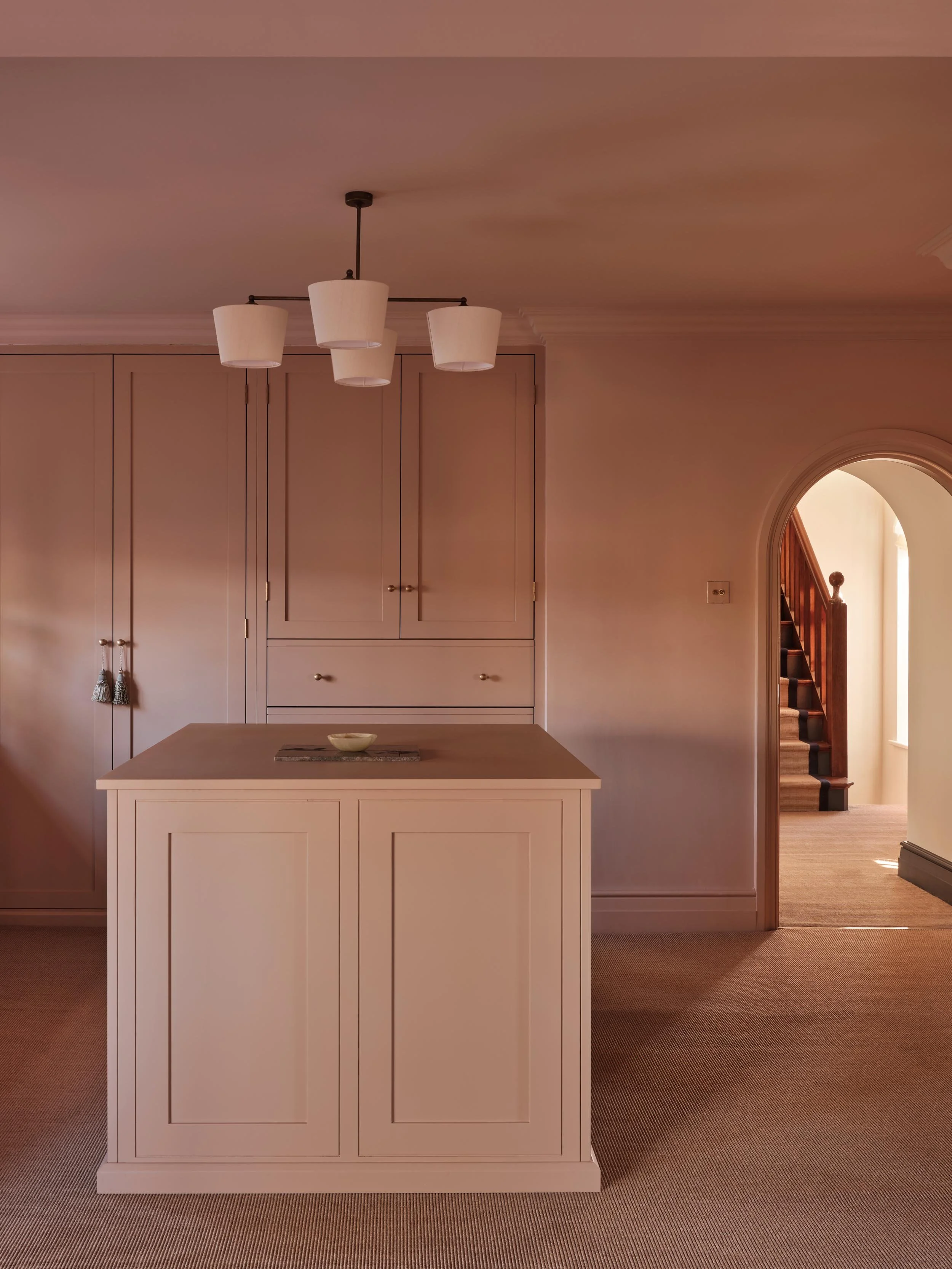 Interior view of a house showing a white kitchen island, beige cabinets, a beige carpeted staircase visible through an arched doorway, and a ceiling light fixture with three white lampshades.