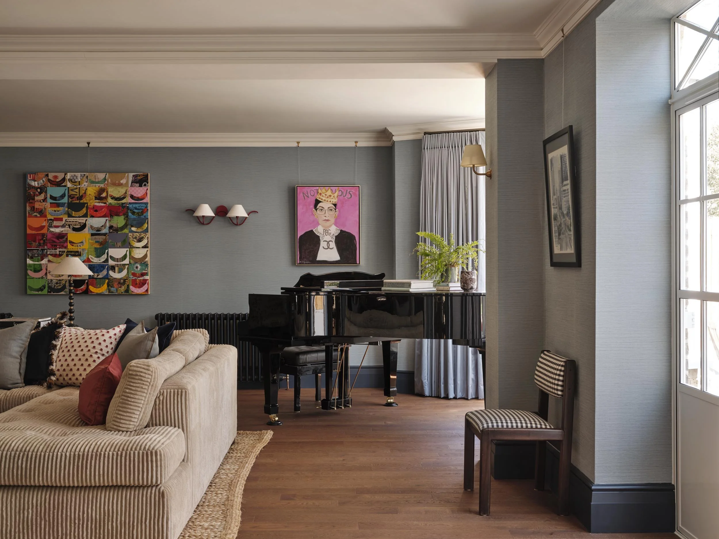 Living room with a beige sofa, pillows, a black grand piano, a colorful modern art piece, a framed portrait with a crown, wall-mounted lamps, window with curtains, and hardwood floors.