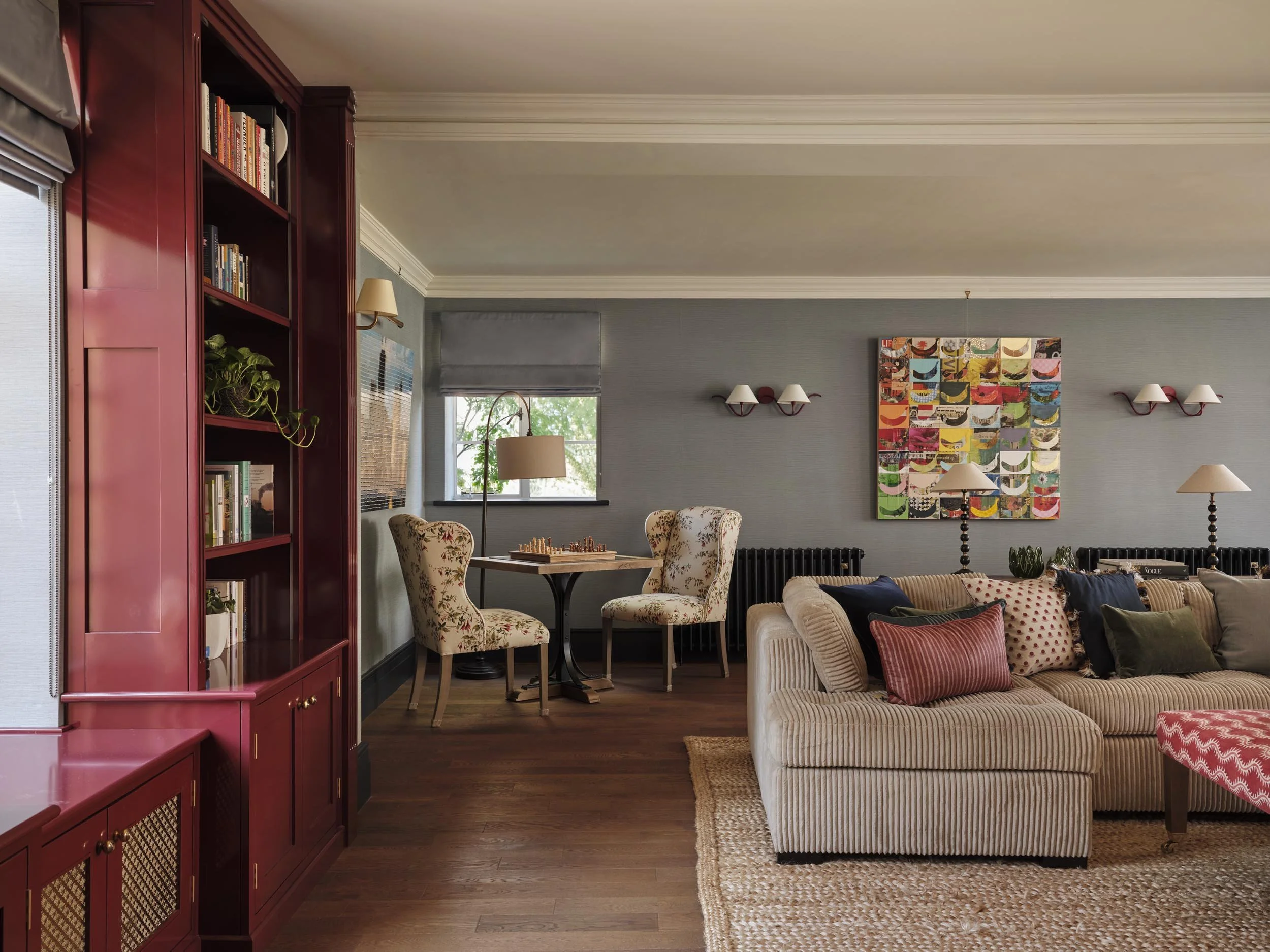 Living room with a red bookshelf, floral armchairs, a beige striped sofa, a colorful abstract wall art, table lamps, and window with blinds.