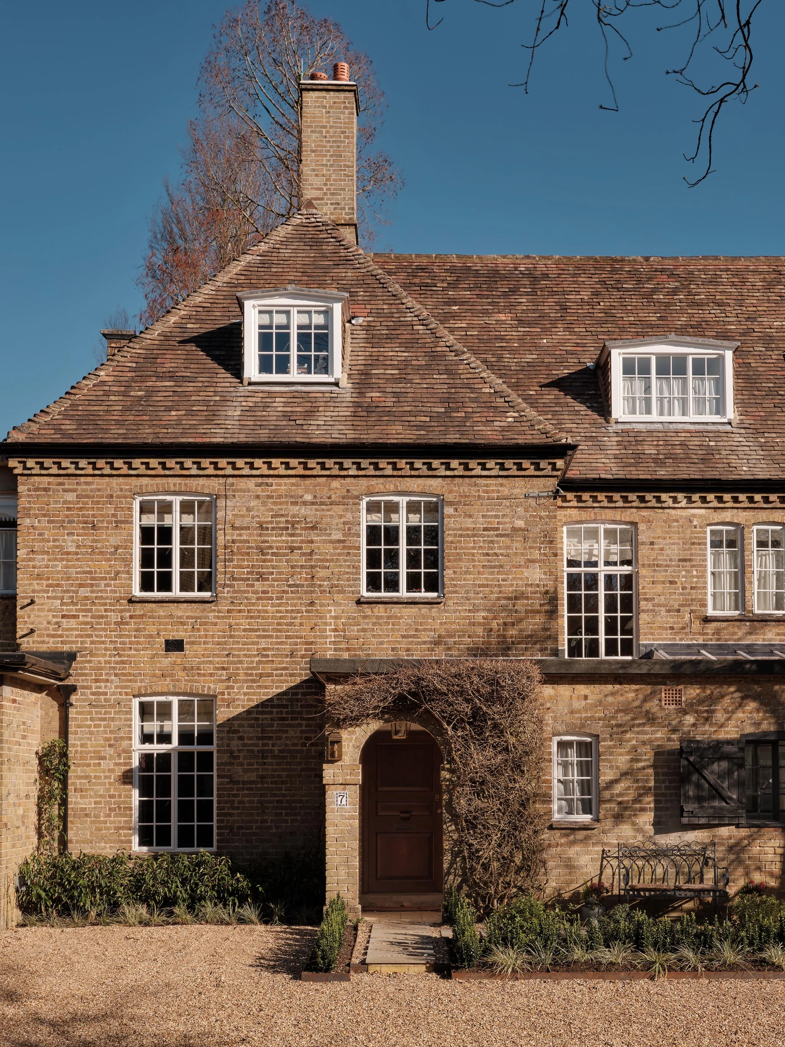 Front view of a traditional brick house with multi-pane windows, a brown front door, a small garden, and a gravel pathway, under a clear blue sky.