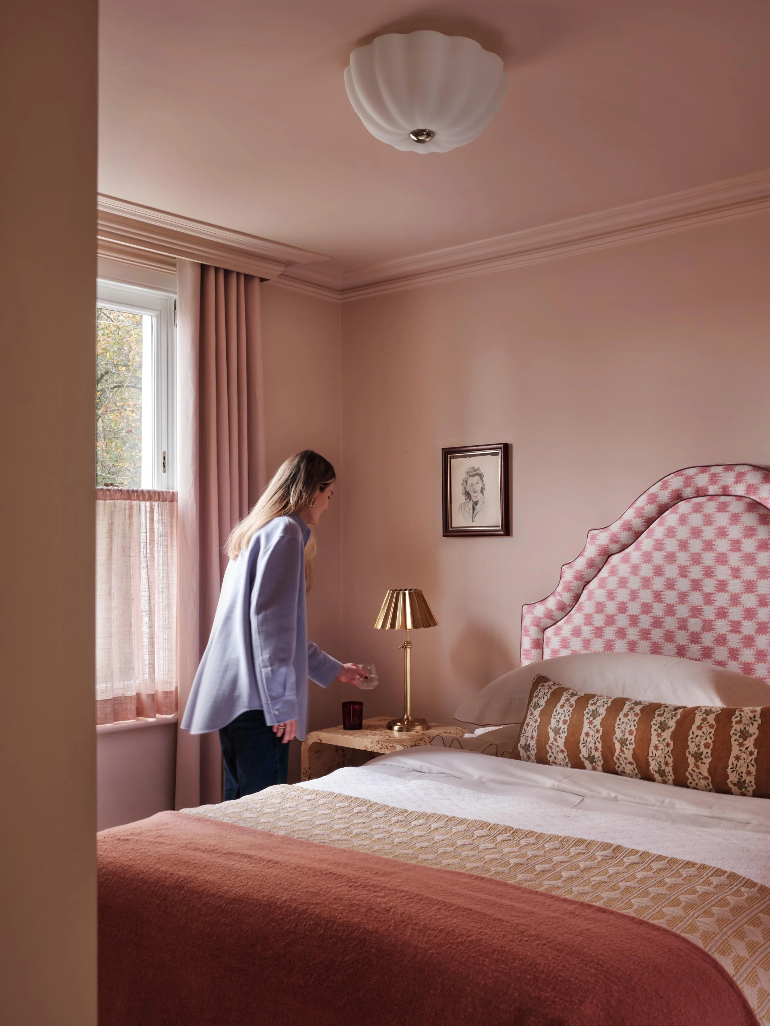 A woman with long hair, wearing a light blue blazer, standing beside a bed in a pink-themed bedroom, with a window and curtains, a framed portrait of a woman on the wall, and a bedside table with a lamp and a glass.