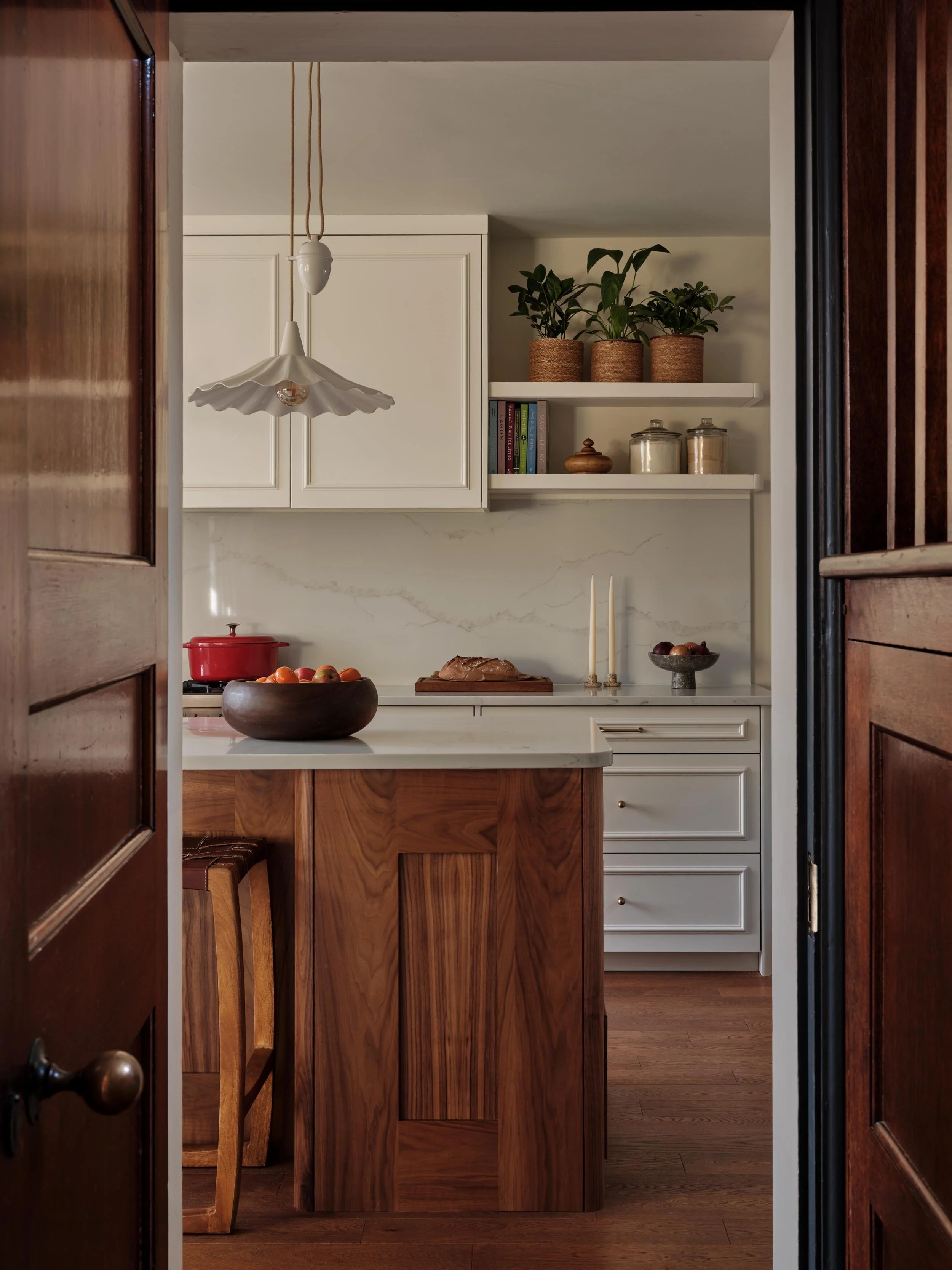View into a kitchen through a partially open wooden door, showing a kitchen island with a wooden base, a bowl of apples, a red pot on the stove, white cabinets, and shelves with decorative items and plants.