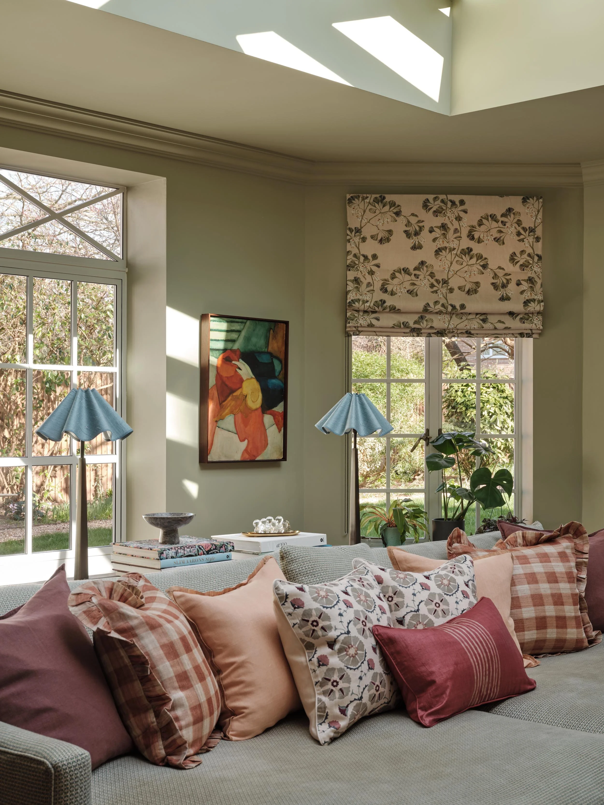 A cozy living room with a large beige sofa adorned with various pink, floral, and plaid throw pillows. Natural light streams through a window with floral patterned Roman shades, and a large painting hangs on the wall. A table in front of the sofa holds a bowl, books, and a decorative tray, with a few potted plants and blue lamps on stands nearby.