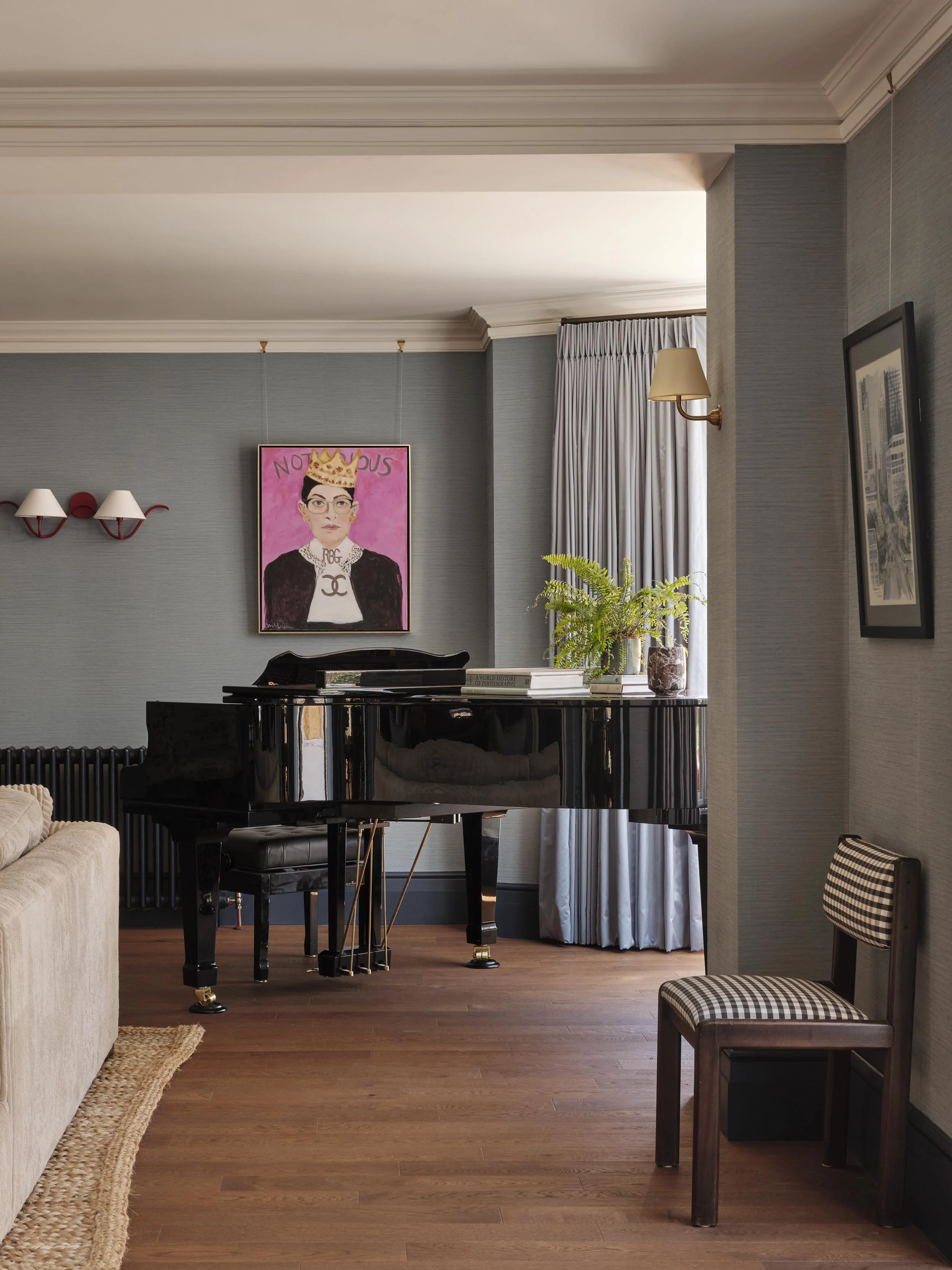 A room with a portrait of a woman wearing glasses, a crown, and a Chanel shirt, hanging on a gray wall. There is a black grand piano beneath the portrait, decorated with books and plants. A wall-mounted lamp is on the right, and a window with white c