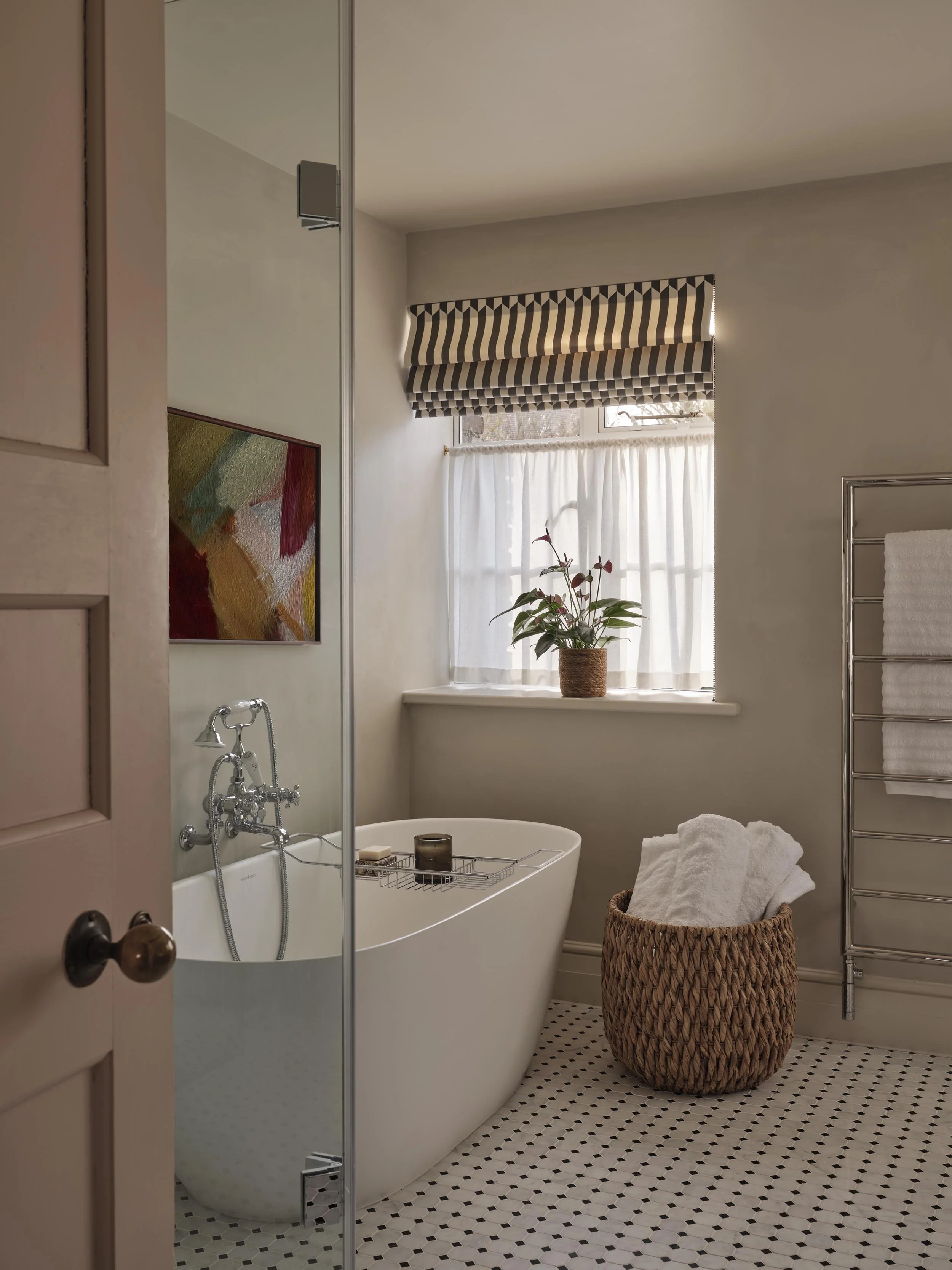 A bathroom with a white clawfoot bathtub, a wicker laundry basket with towels, a potted plant on the windowsill, a patterned valance over the window, a towel rack with white towels, and a partial view of a shower with glass door.