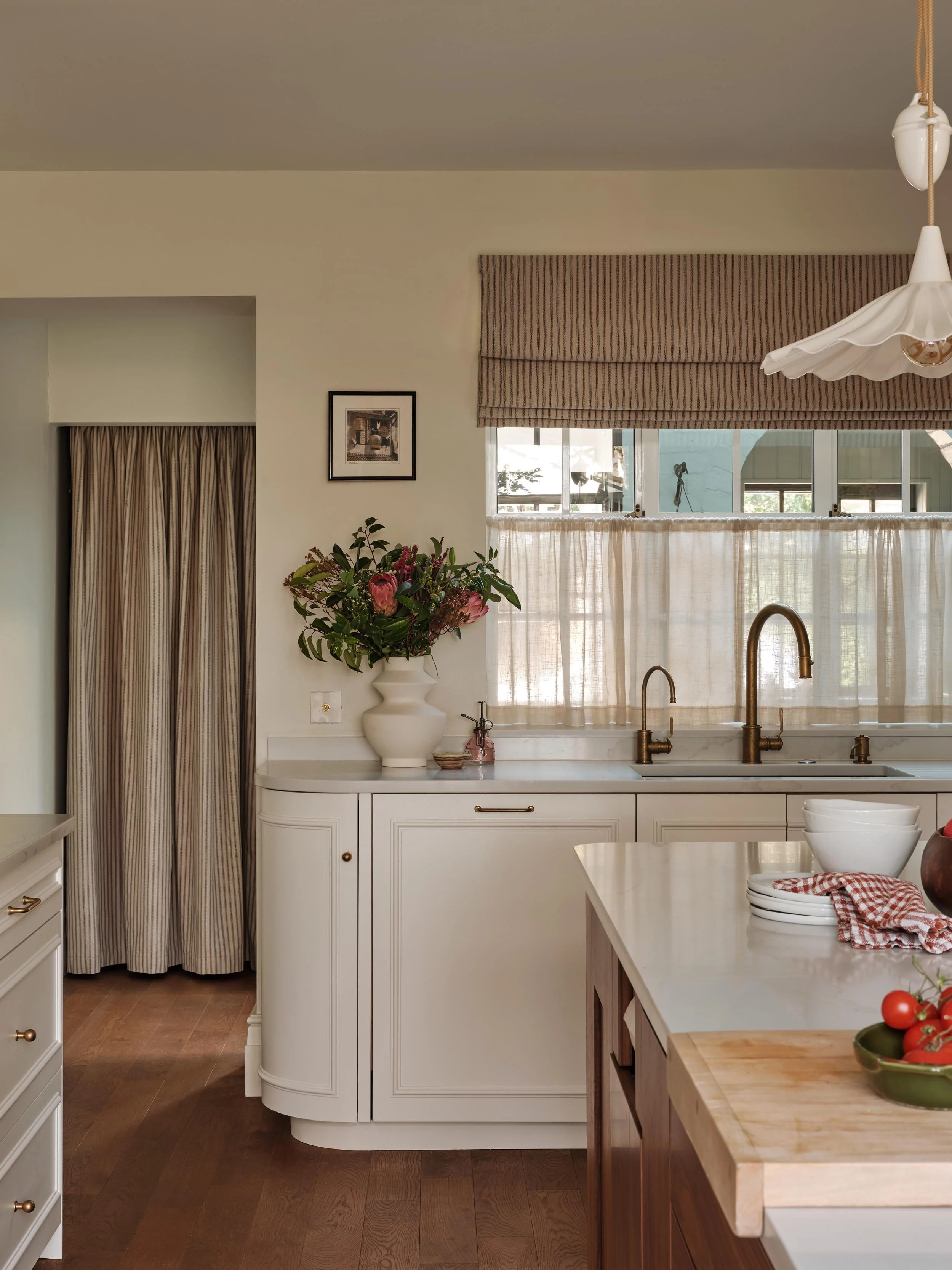 A kitchen with a beige countertop, gold faucet, white cabinetry, gold handles, a window with beige striped curtains, a vase with pink and purple flowers, and a wooden cutting board with white bowls and a red and white checkered cloth.
