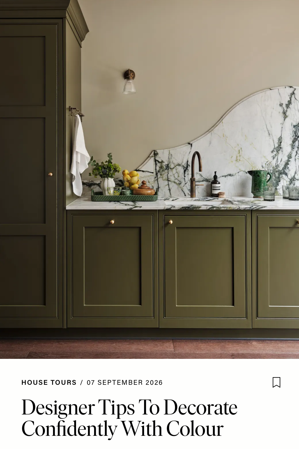 A kitchen with green cabinets, a marble countertop, and a marble backsplash. The countertop holds a green tray with lemons, a small potted plant, and a glass bowl. There is a black soap dispenser, a green mug, and drinking glasses on the right side near the sink with a copper faucet. A towel hangs from the cabinet on the left, and a wall sconce is above.