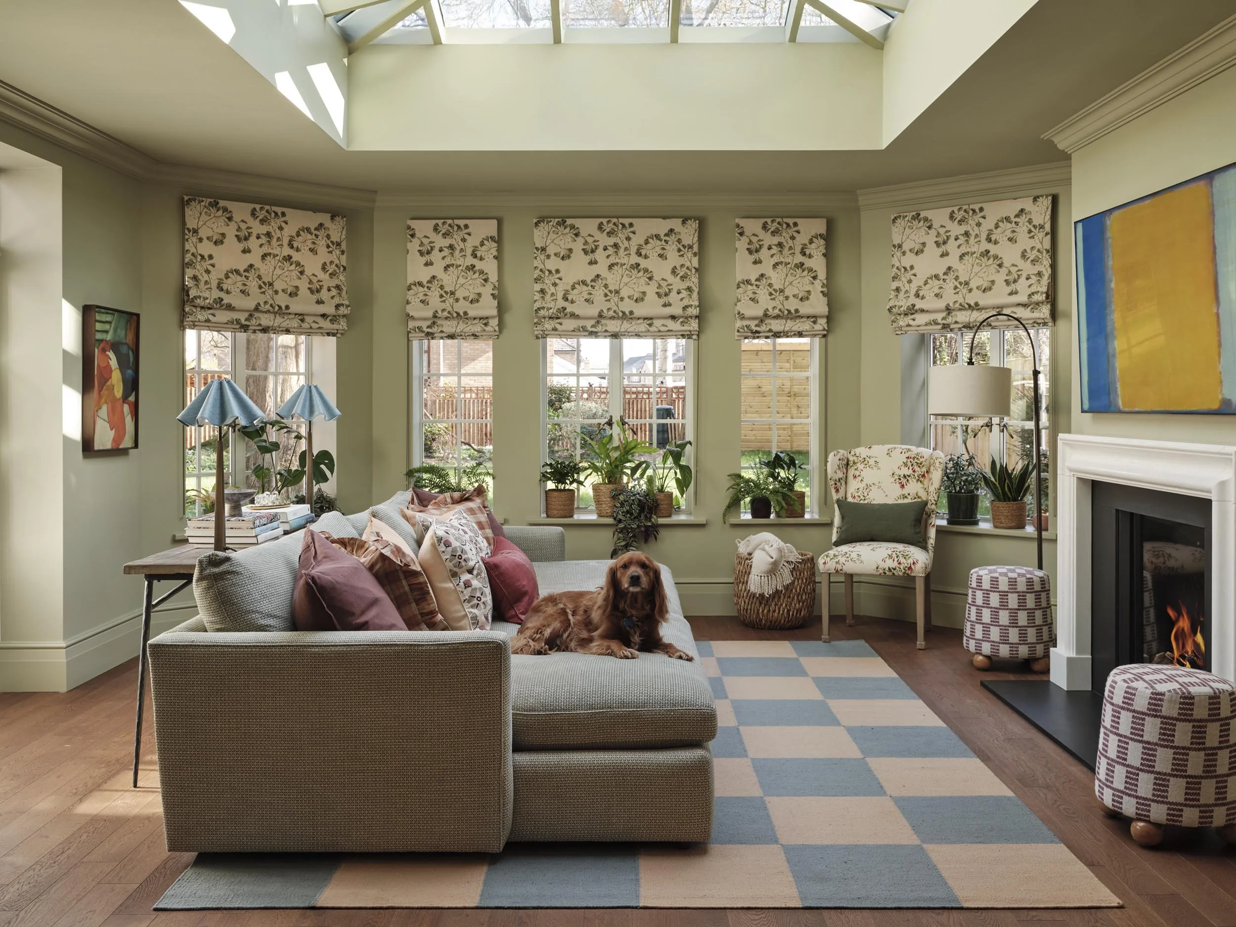 Bright living room with large windows, green walls, plants, colorful artwork, a couch with pillows, a side table with blue lamps, a cozy armchair near a fireplace, and a dog sitting on the couch under a skylight.