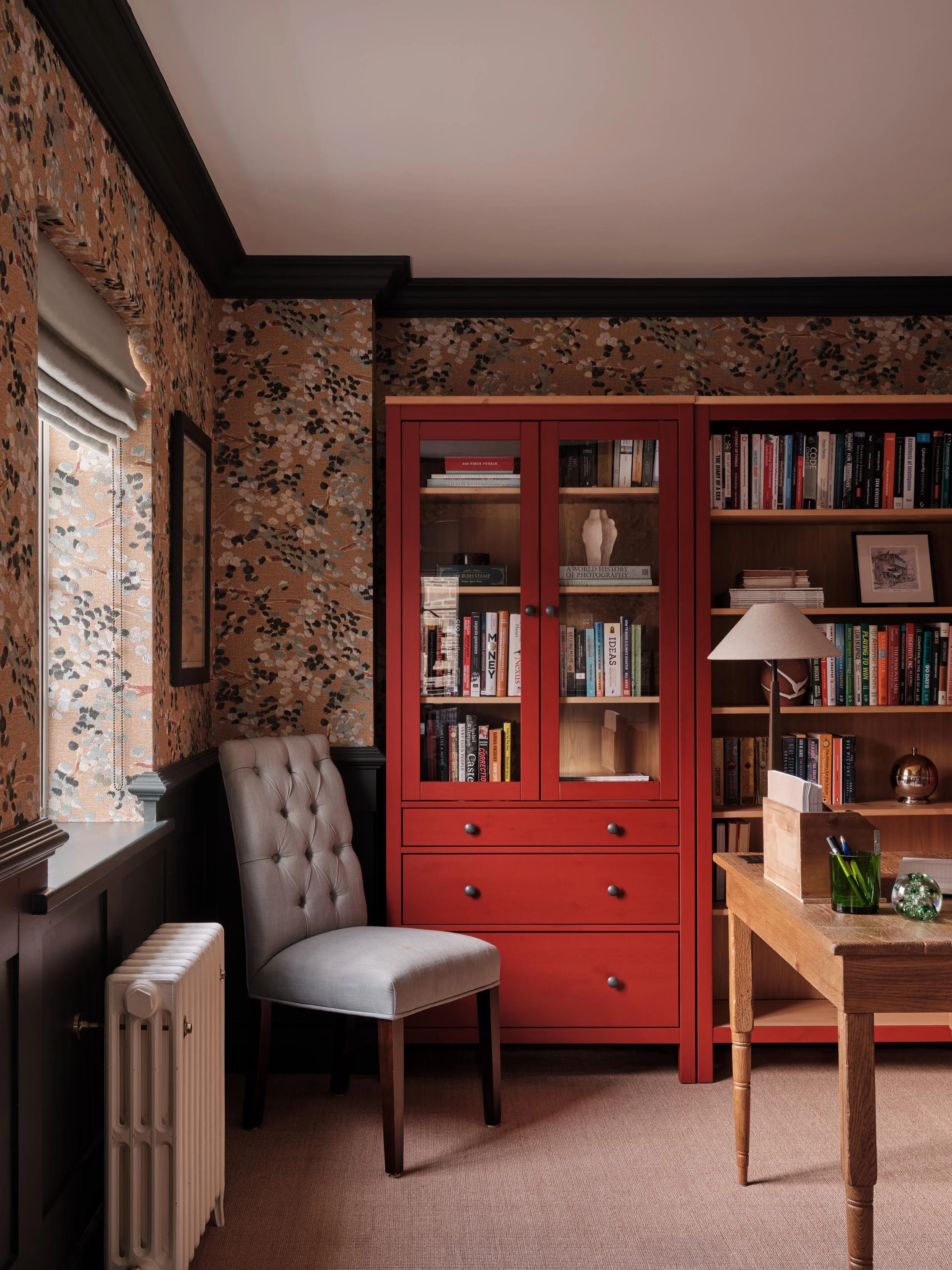 A cozy reading nook with a tufted beige armchair next to a window with Roman shades, a red bookshelf filled with books and decorative items, a wooden desk with green and glass decor, a table lamp, and a patterned wallpaper in shades of brown, black, 