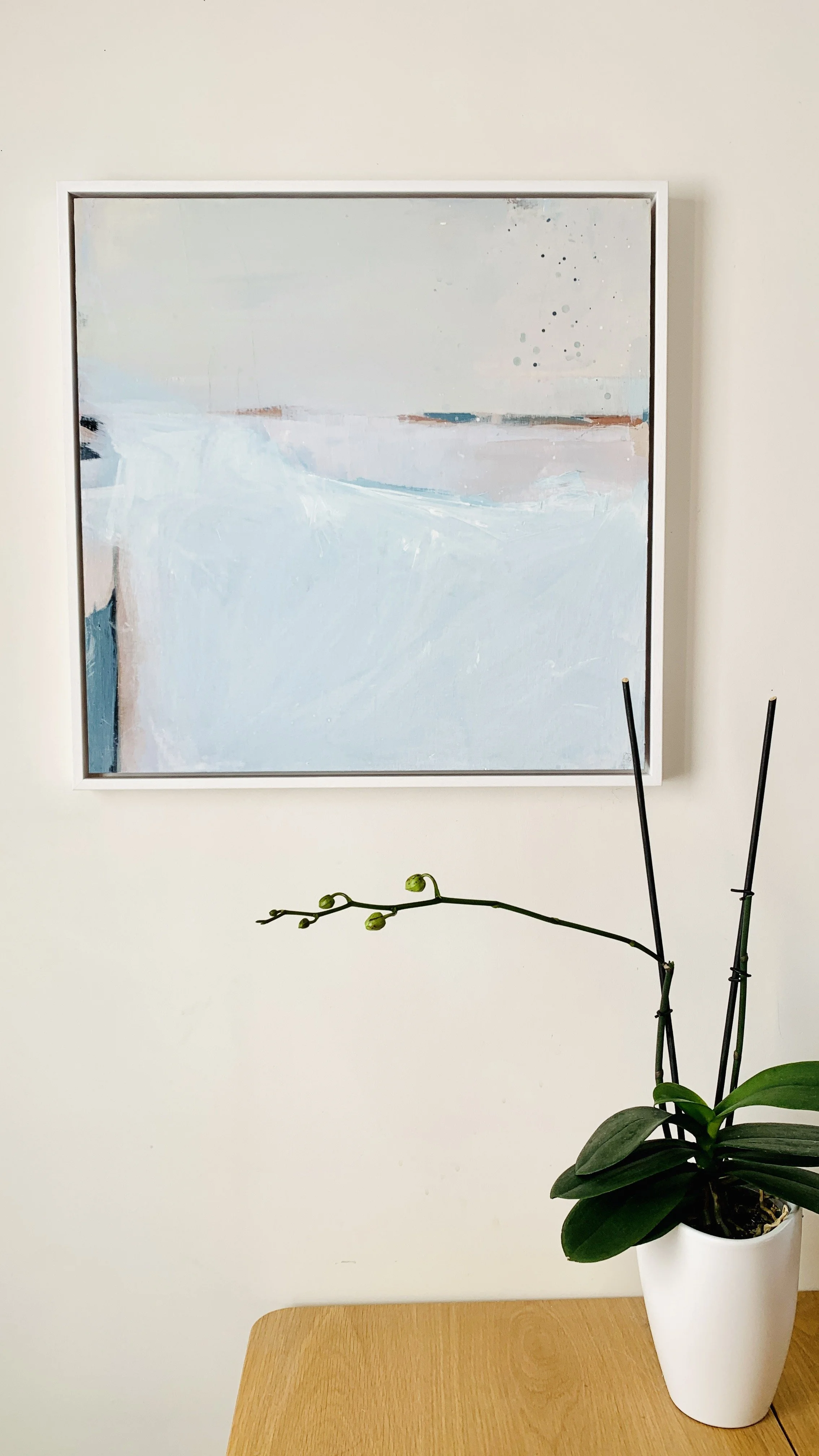 A white potted orchid with green leaves and budding stems on a wooden surface, with a framed abstract painting in soft pastel colors on a white wall behind it.