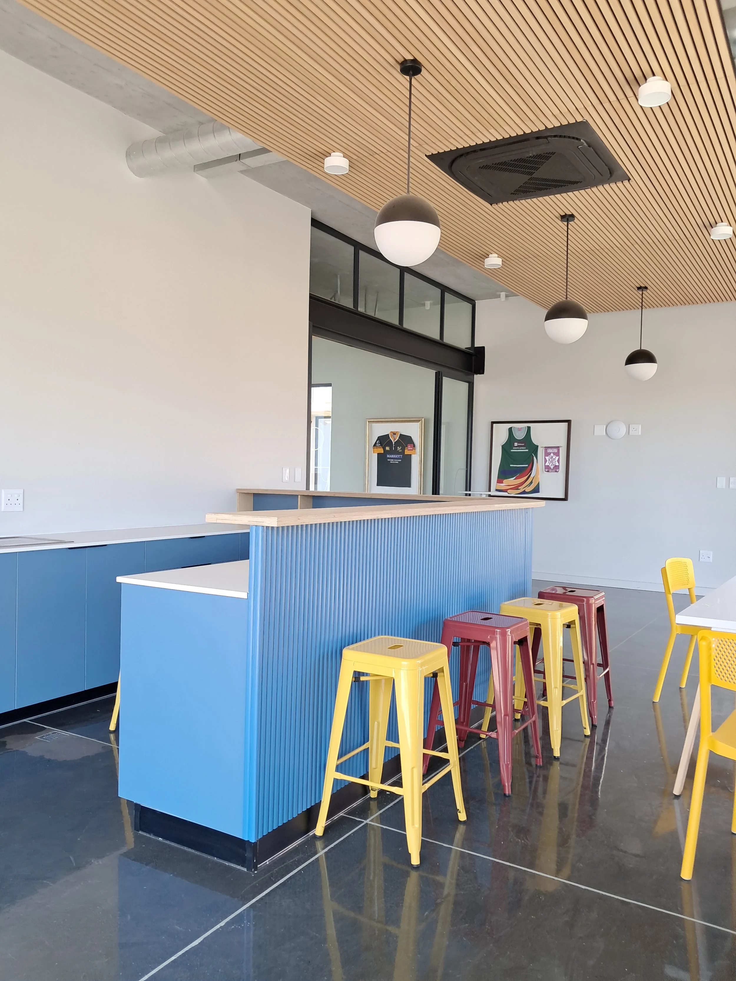 Modern interior space with a wooden ceiling, hanging round black and white pendant lights, colorful stools, flueted wood  painted and granite counter, framed sports jerseys on the wall, and black tiled flooring.