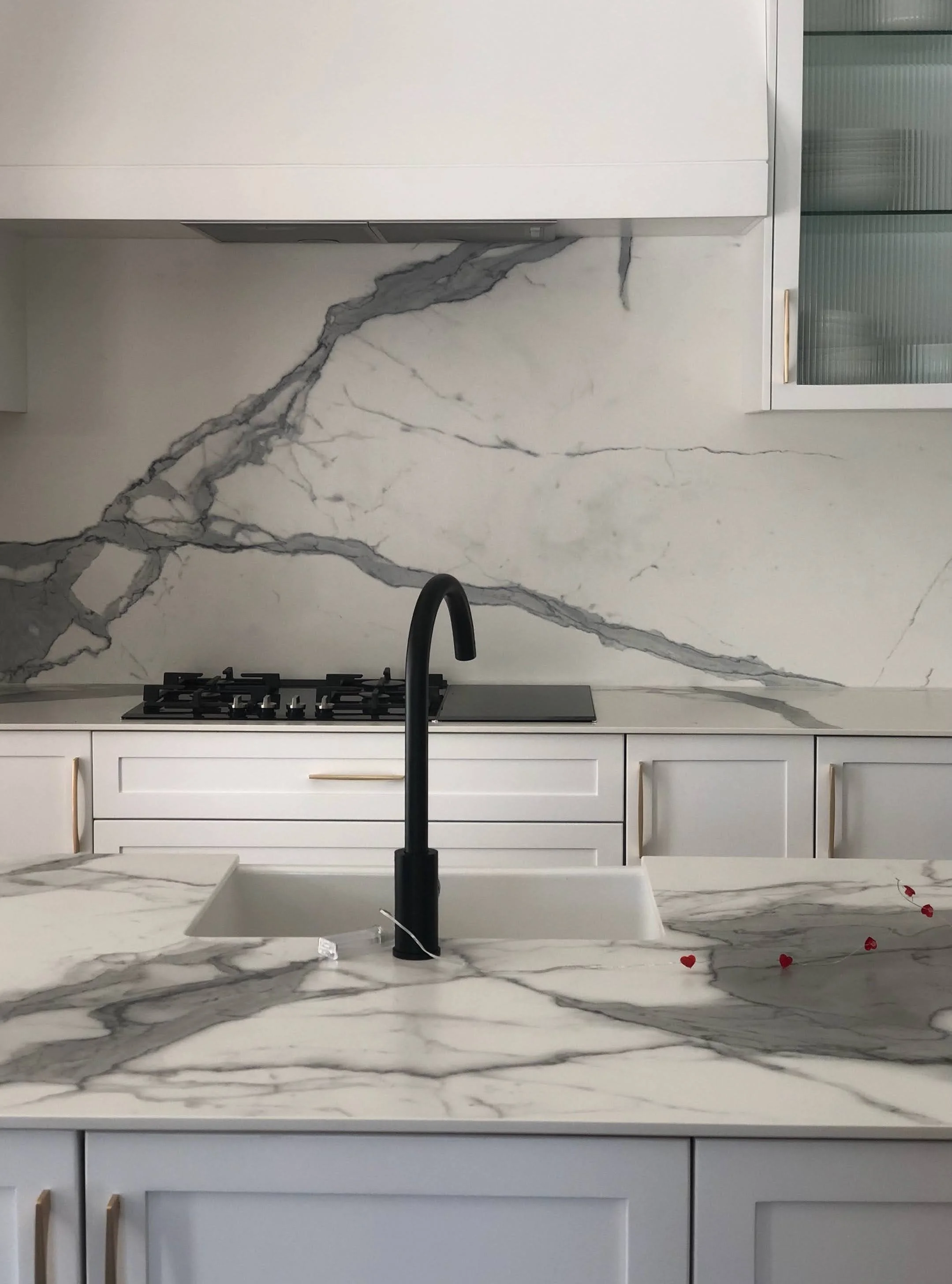 A classic contemporary kitchen with white cabinetry, a marble backsplash with gray veining, a black tap, a white square sink, a stove, and glass-front cabinets. Heart-shaped decorations on the granite countertop.