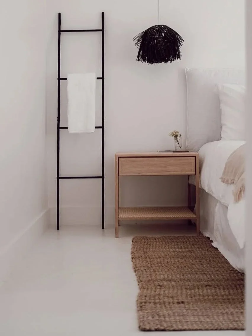 A bedroom corner with a white wall, a wooden nightstand, a small plant on it, a bed with white bedding, a black hanging lamp, a black ladder-style towel rack with a white towel, and a woven jute rug on the white floor.