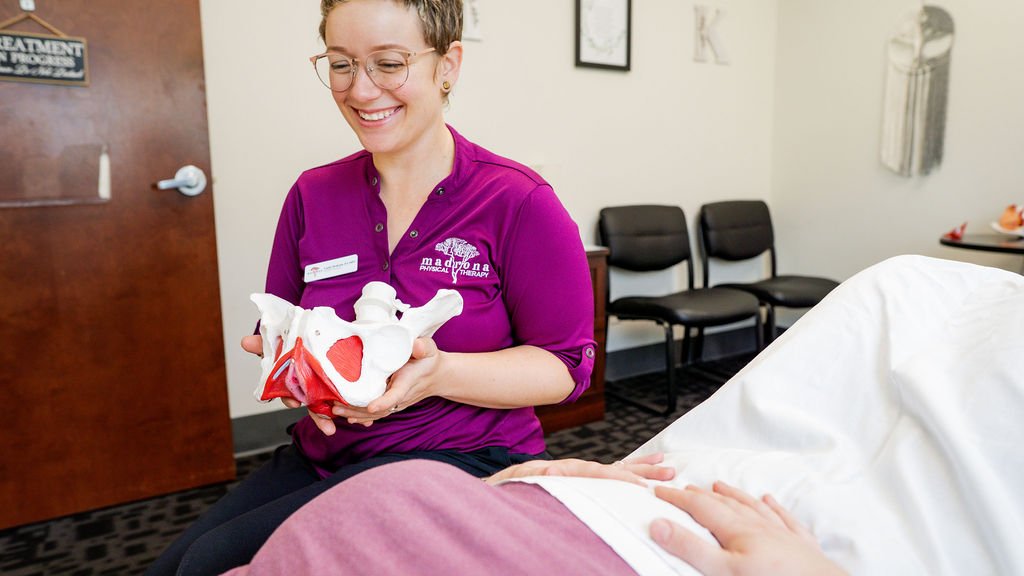Pelvic floor physical therapy provider smiling warmly at reclined patient, showing female pelvic model.