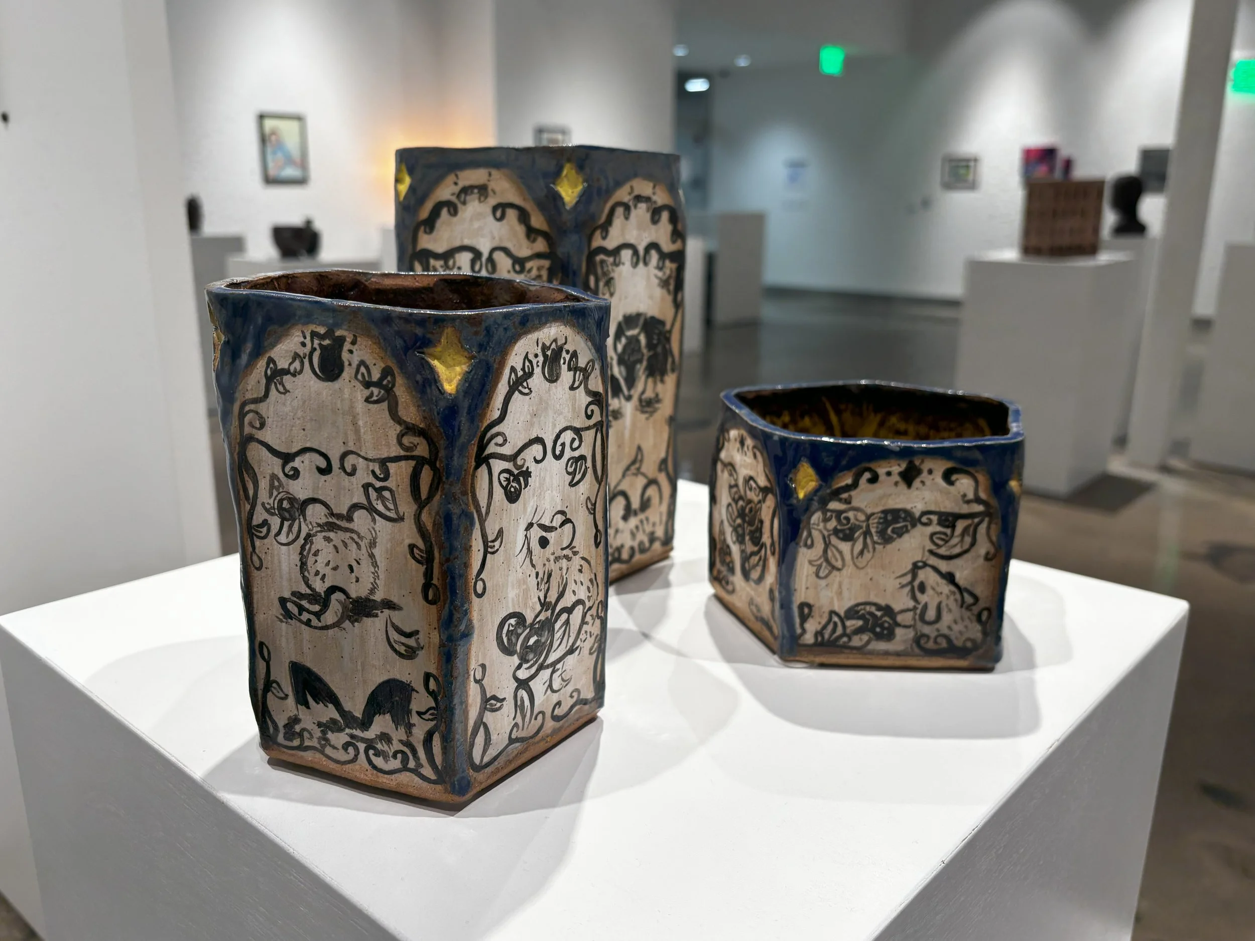  Three hand-built ceramic vessels on a white pedestal, white-glazed surfaces with illustrations of woodland animals and leaves. 