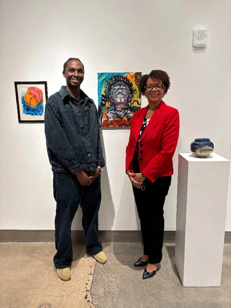  A young man and a woman smiling side-by-side next to displayed paintings. 